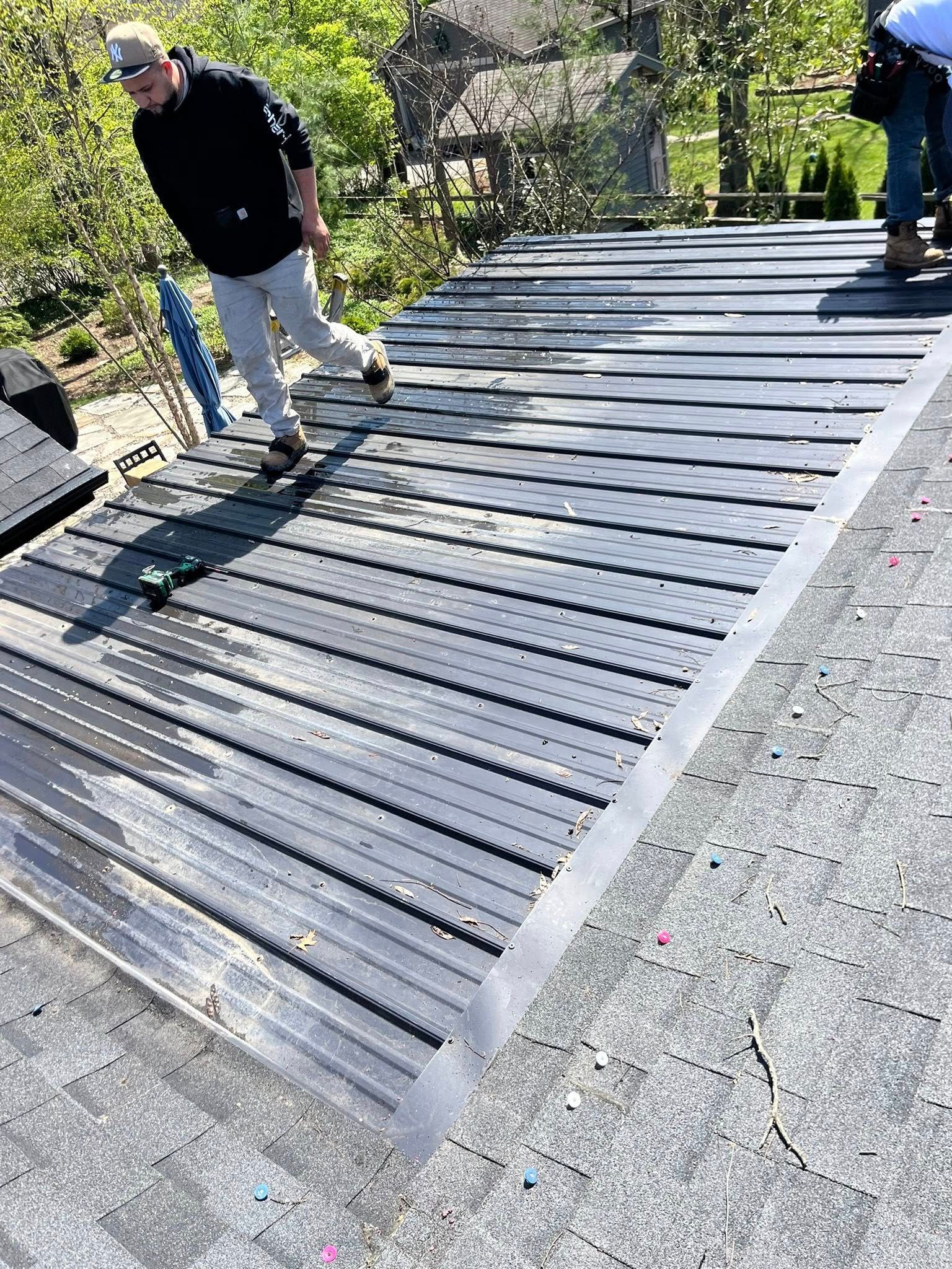 A person in a black hoodie and light pants stands on a sloped roof installing rows of black, ridged roof panels.