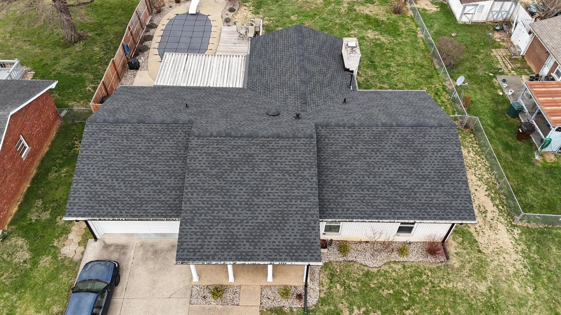 An aerial view of a residential house with a dark grey shingled roof, a front porch, and a backyard with a pool cover.