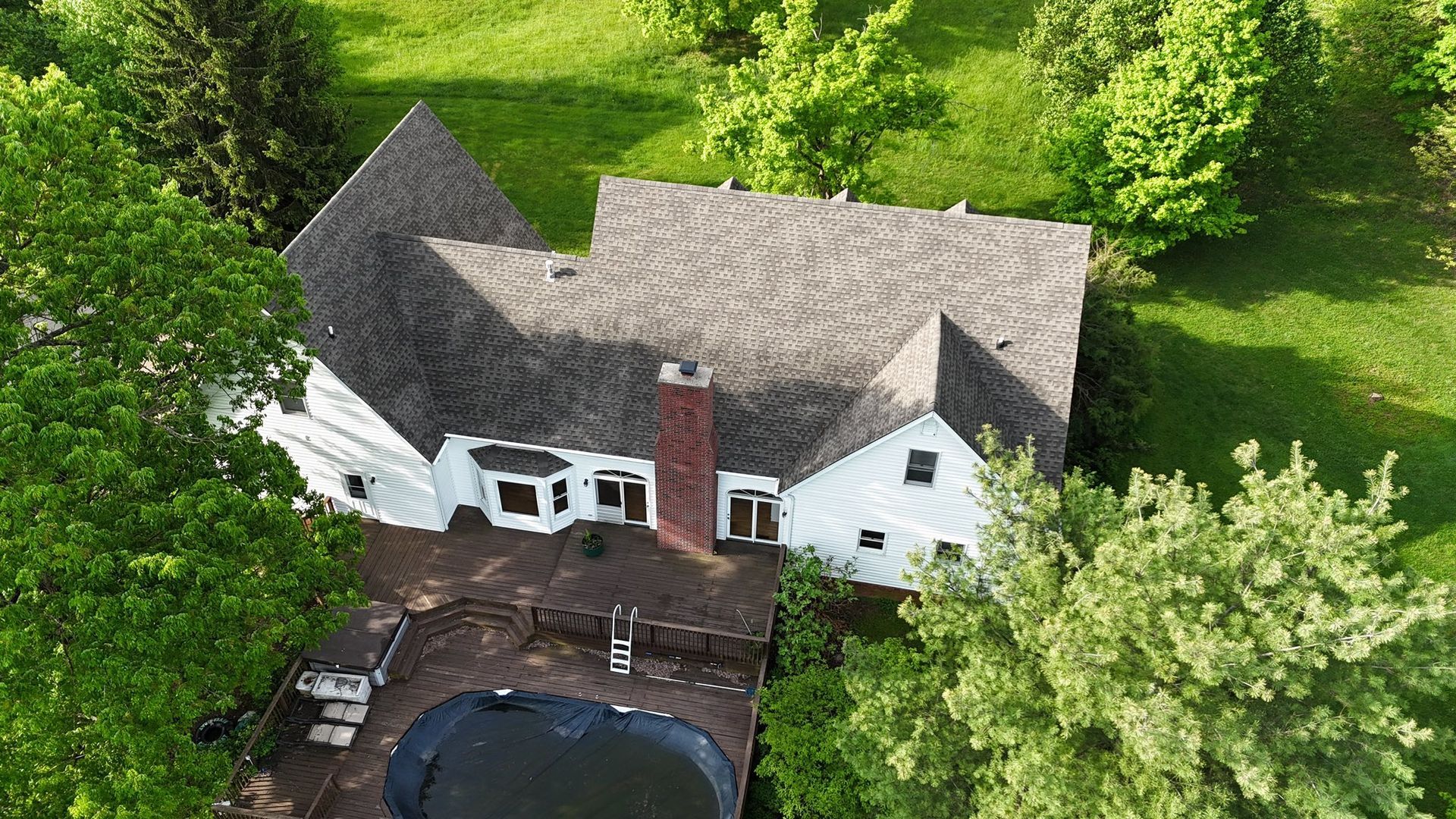 Aerial view of a white, multi-roofed house with a red brick chimney and a backyard swimming pool surrounded by trees.