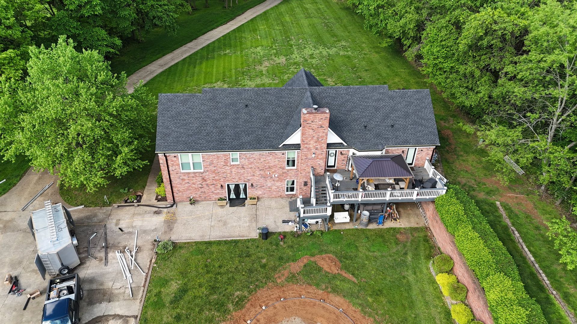 Aerial view of a two-story brick house with a dark shingled roof, a back deck, and a yard in a wooded suburban setting.