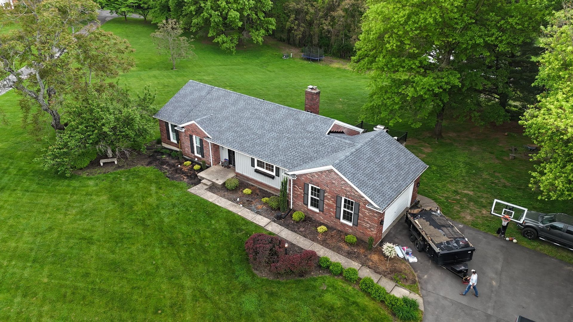 Aerial view of a brick house with a new grey roof, showing a dumpster trailer in the driveway and two people working.