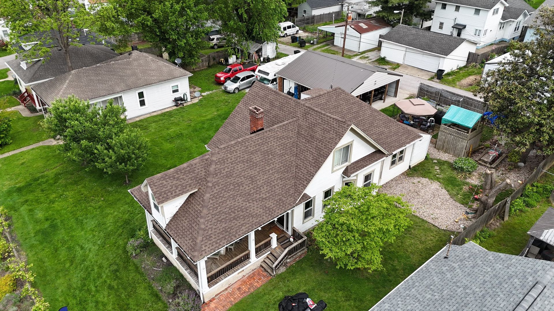 Aerial view of a white house with a brown shingled roof, a covered porch, and surrounding green lawn in a neighborhood.
