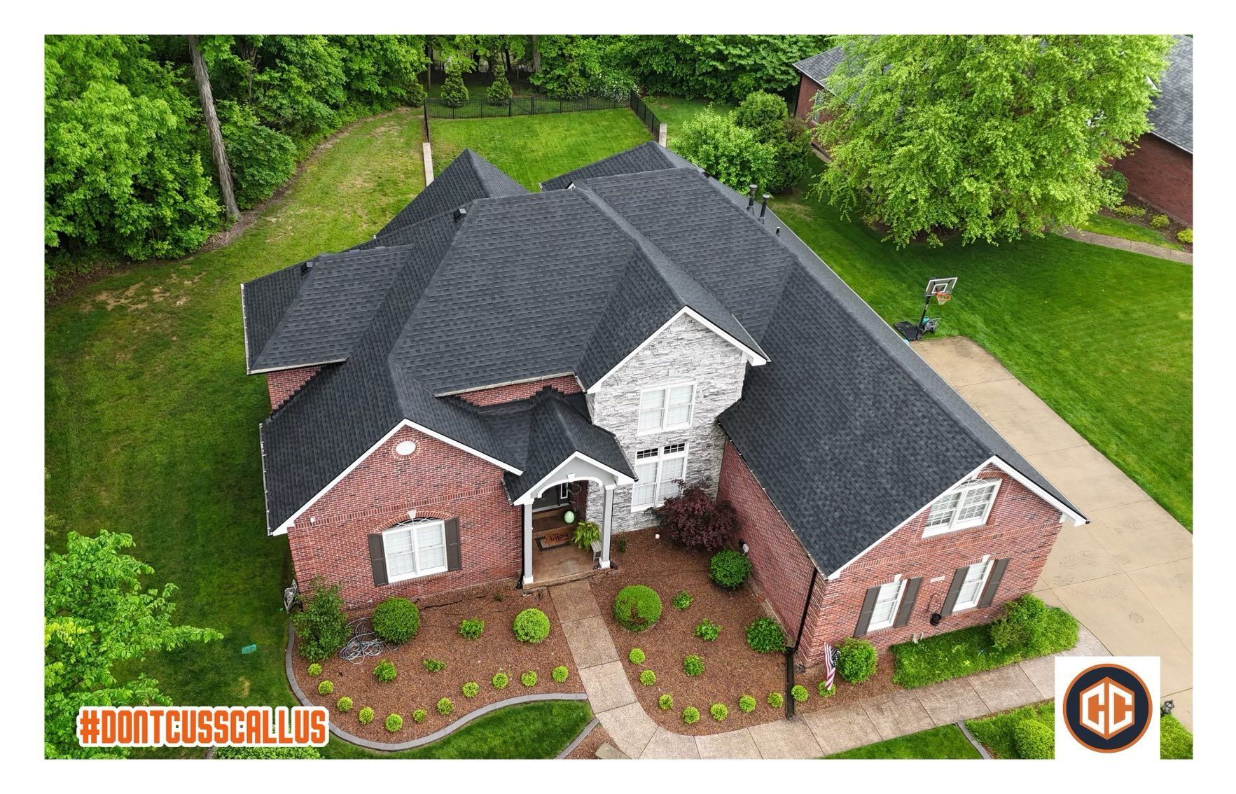 An aerial view of a single-story brick house with a dark shingled roof, surrounded by a green lawn and trees.