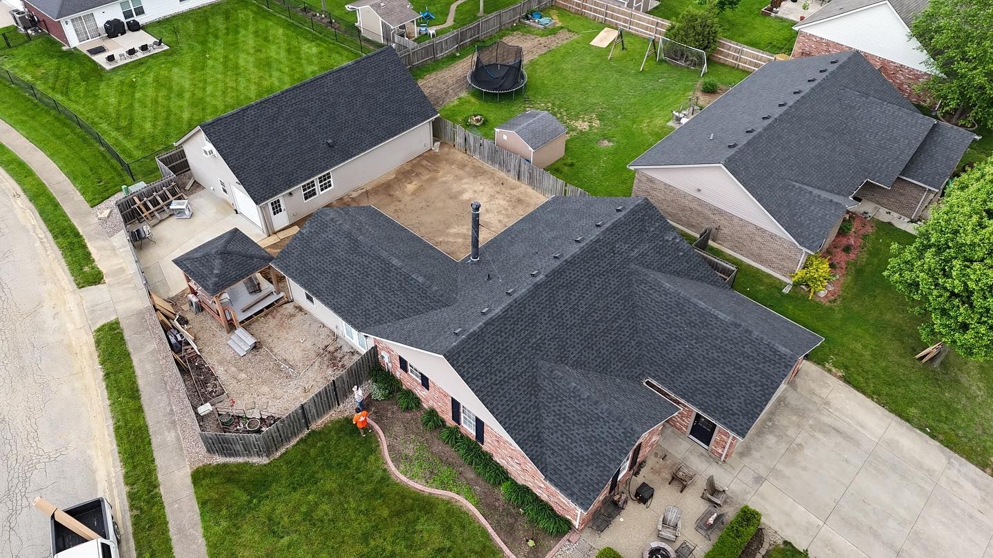 Aerial view of a residential suburban house with dark gray shingles, a patio, and a partially landscaped backyard.
