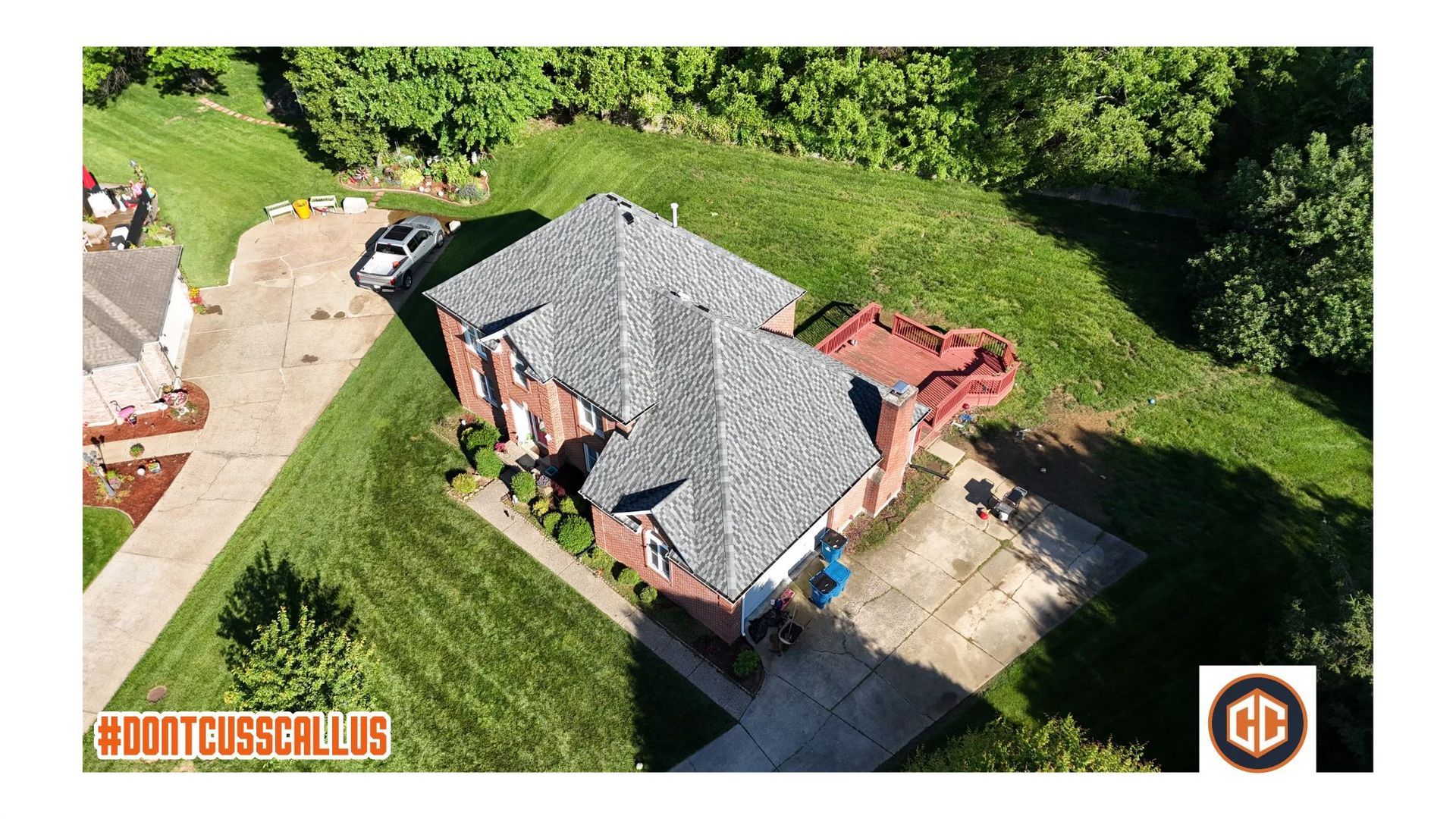An aerial view of a brick house with a gray shingled roof, surrounded by a lawn, driveway, and a red deck.