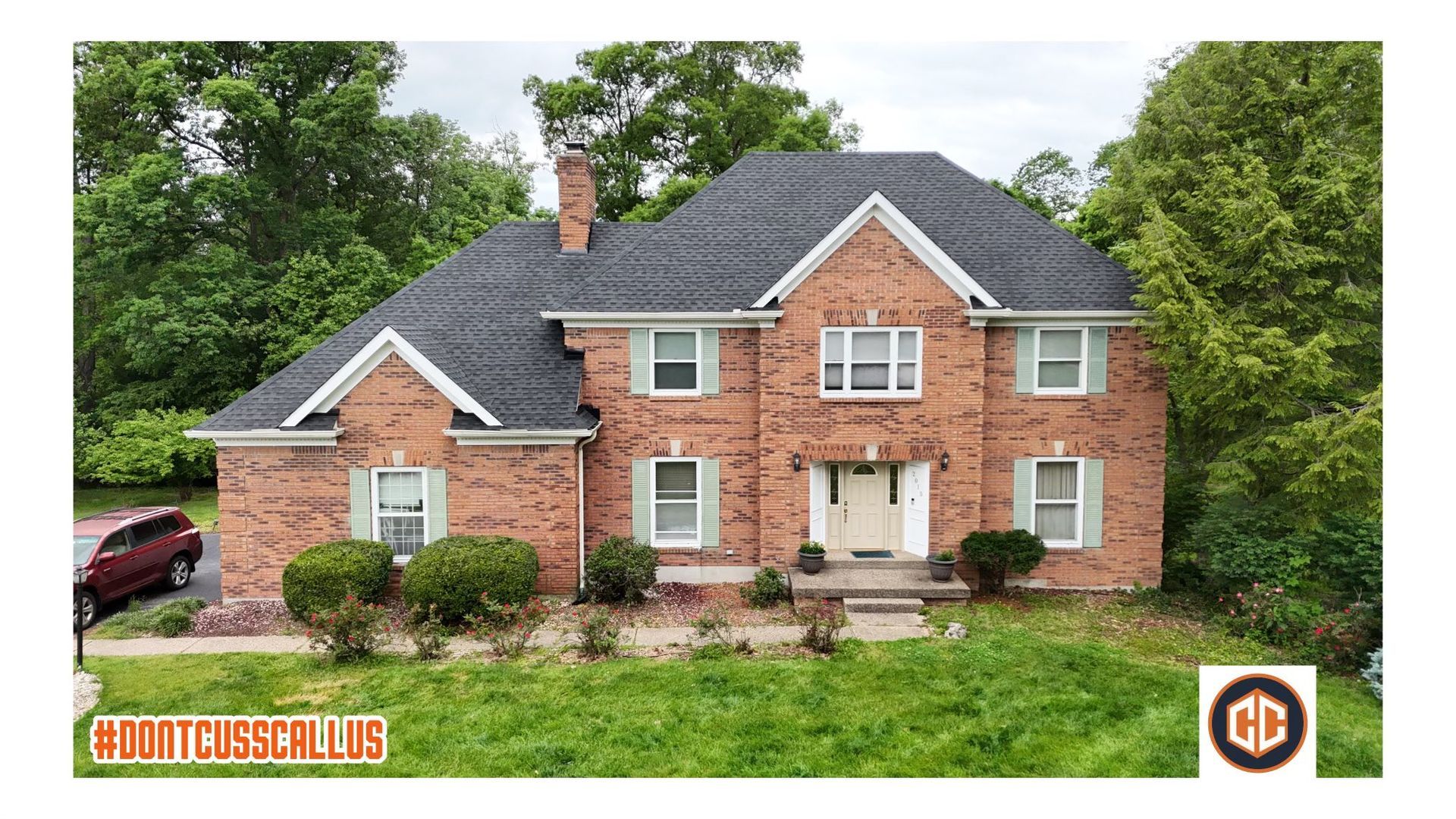 A two-story brick house with a dark shingled roof, light-colored shutters, and a front lawn, framed by lush green trees.