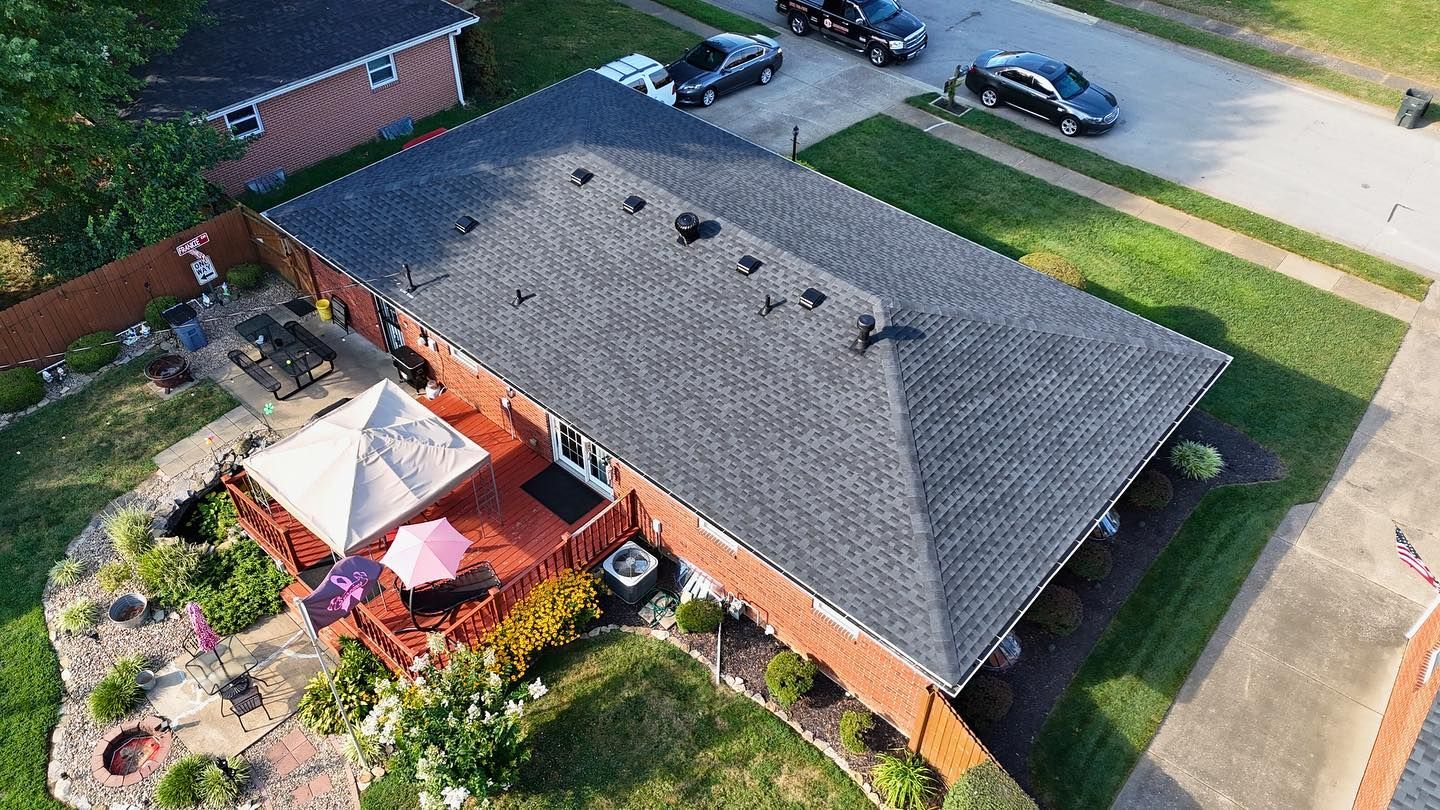 Aerial view of a brick house with a dark shingled roof, a wooden deck with umbrellas, and a backyard patio and lawn.