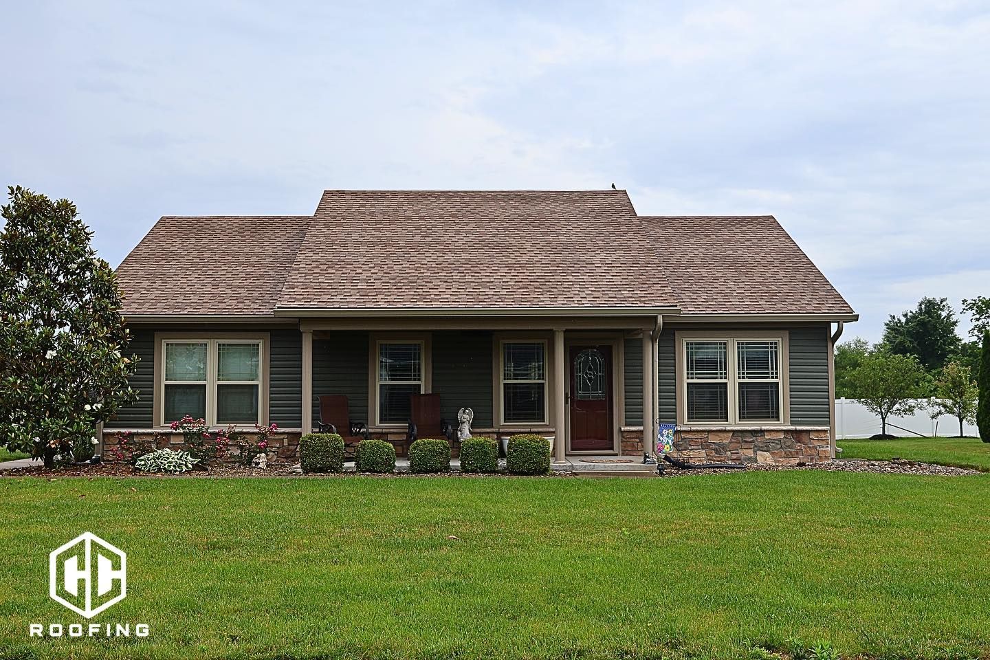 A single-story house with a brown shingled roof, dark horizontal siding, and a brick base under a clear blue sky.