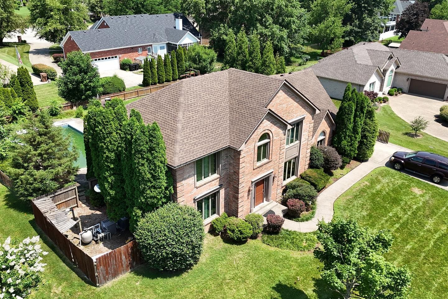 Aerial view of a brick house with a brown shingled roof, surrounding green lawn, trees, and a backyard pool.