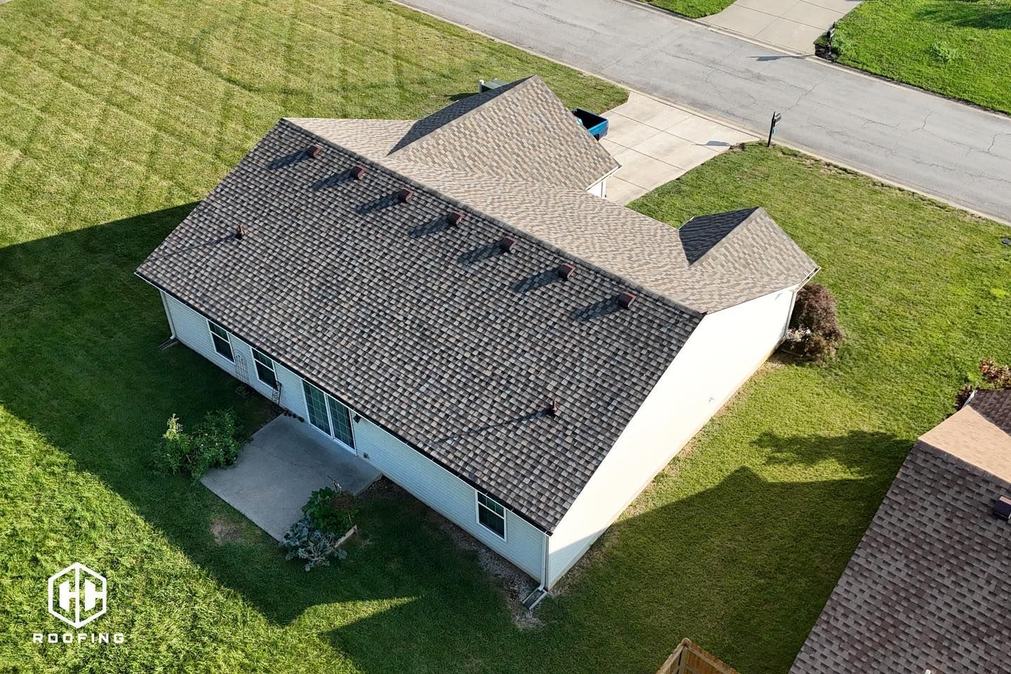 Aerial view of a residential house with a grey shingled roof, white siding, and a concrete patio, set in a green yard.