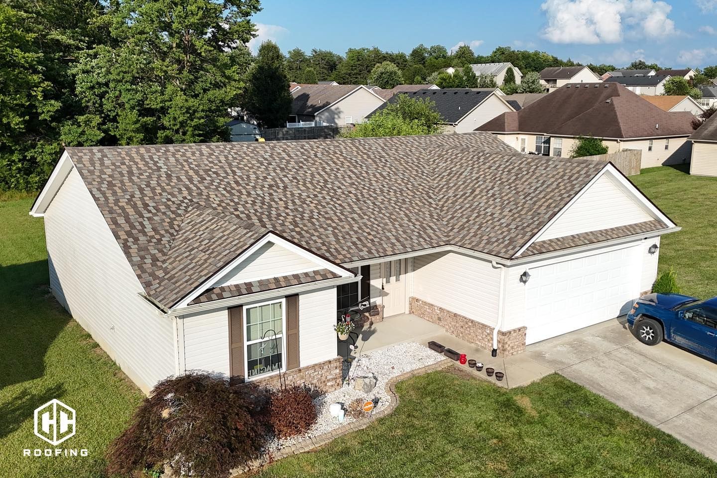 An aerial view of a single-story, beige residential house with a shingled roof, front porch, and a blue car in the driveway.