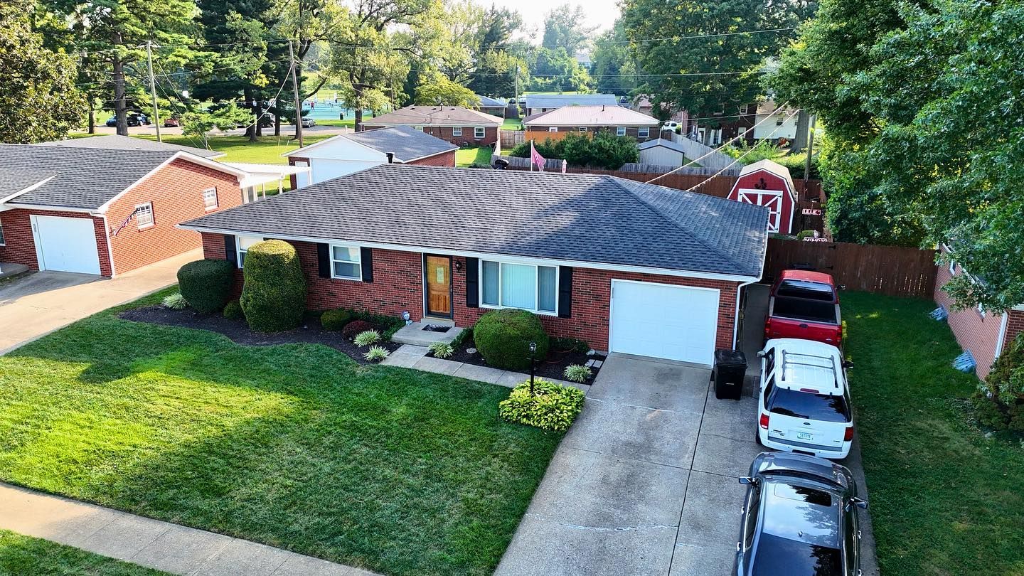 Aerial view of a single-story brick house with a dark shingled roof, a green lawn, and three parked cars in the driveway.