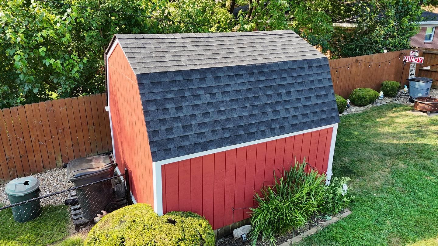 A red wooden shed with a two-toned gray shingled roof sits in a backyard next to a wooden fence and green landscaping.