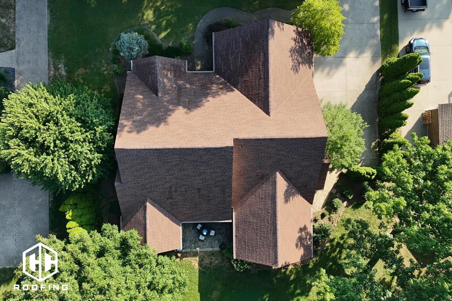 A top-down aerial view of a house with a complex brown shingled roof, surrounded by green trees and a paved driveway.