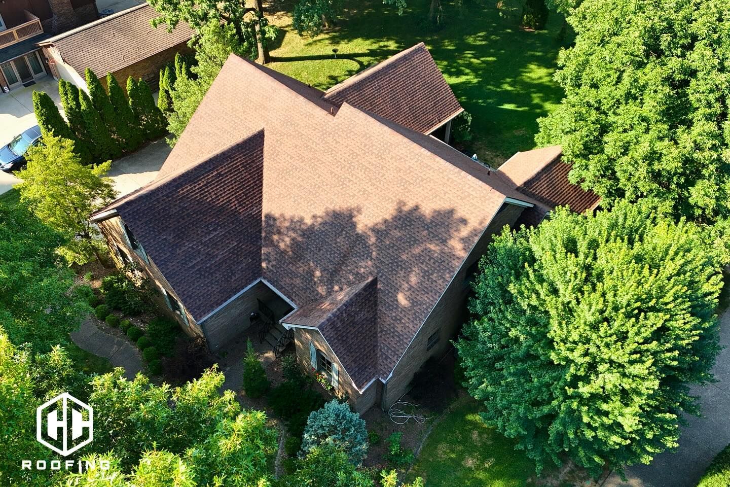 Aerial view of a residential house with a dark brown shingled roof, surrounded by lush green trees.