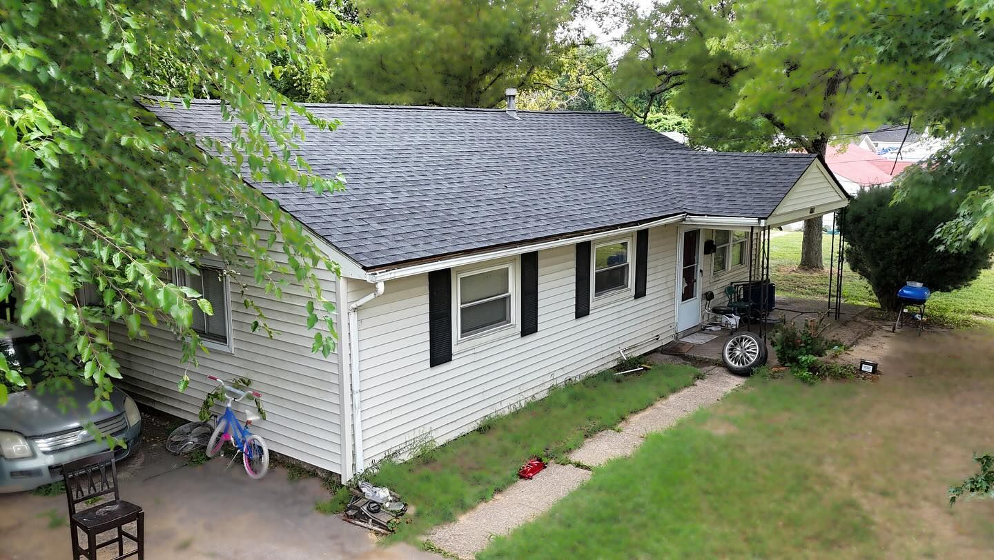 A white, single-story house with a grey roof and black shutters sits on a grassy lot with trees and a parked car nearby.