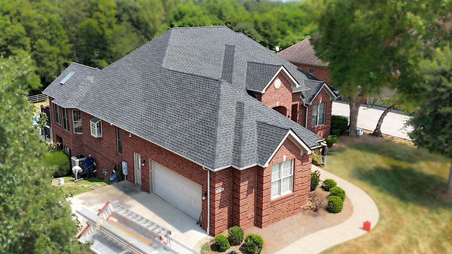 An aerial view of a two-story red brick house with a dark grey shingled roof, surrounded by trees and a concrete path.