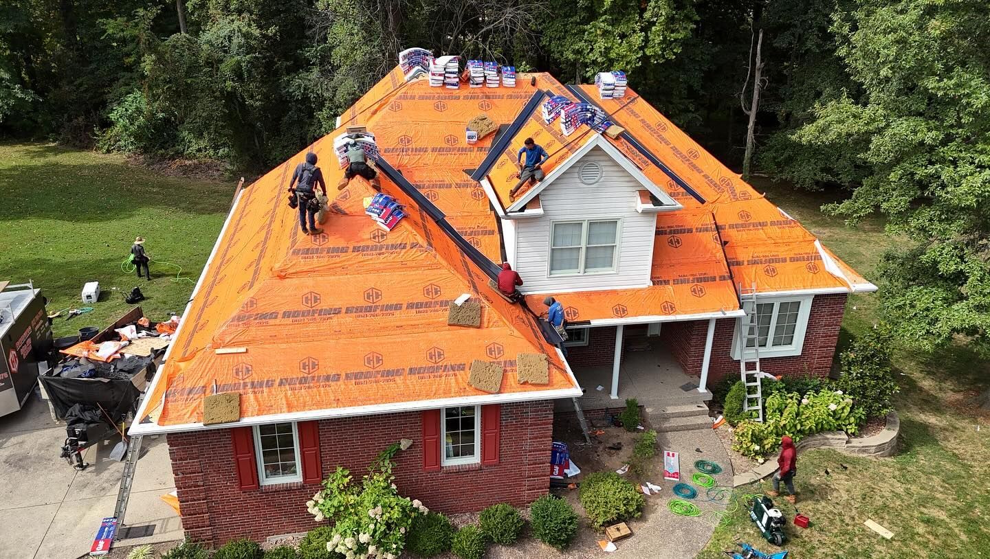 Aerial view of a crew installing bright orange roofing underlayment on a brick house during a residential renovation.