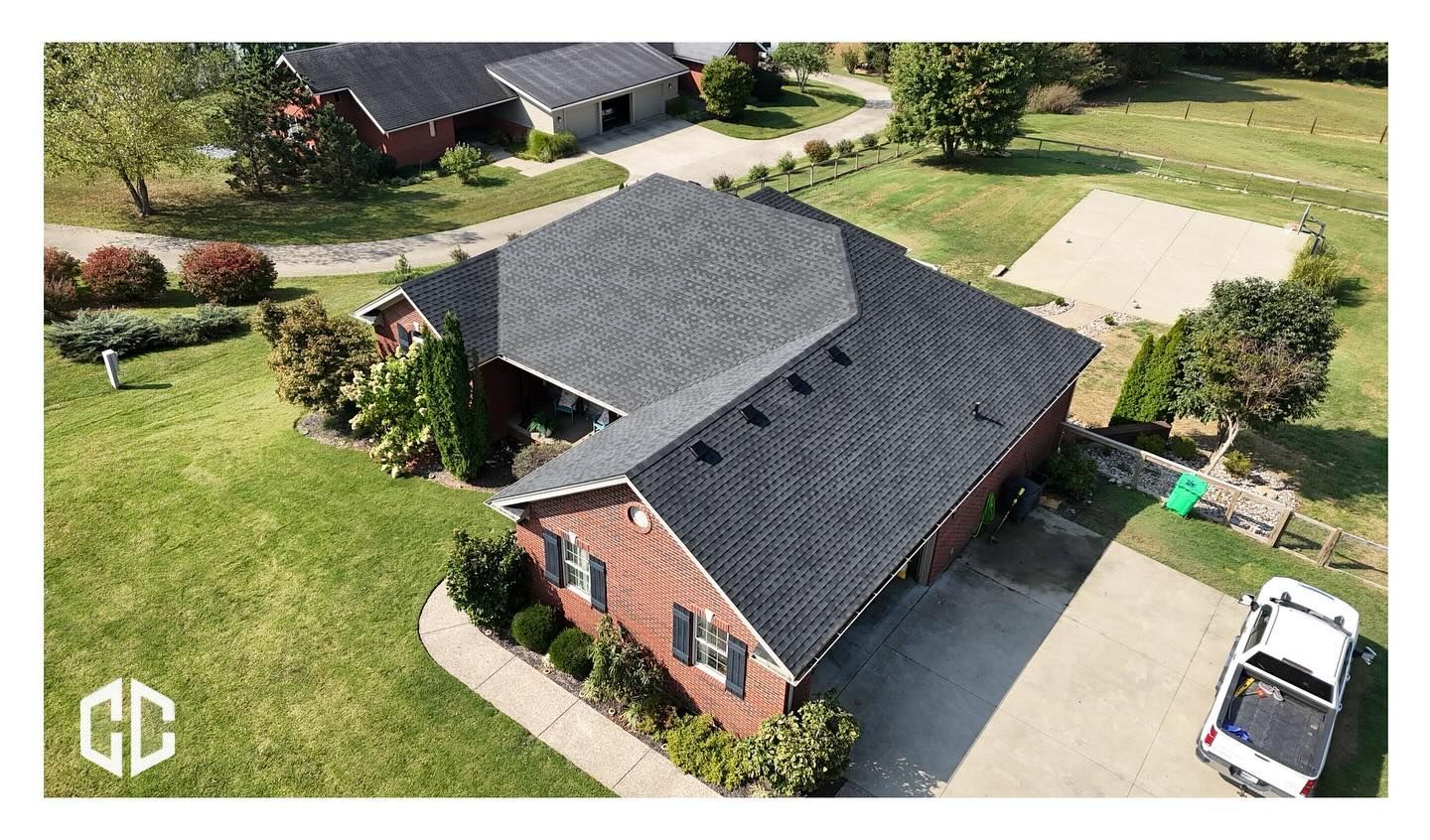 Aerial view of a red brick house with a dark asphalt shingle roof, surrounded by a green lawn and a driveway.