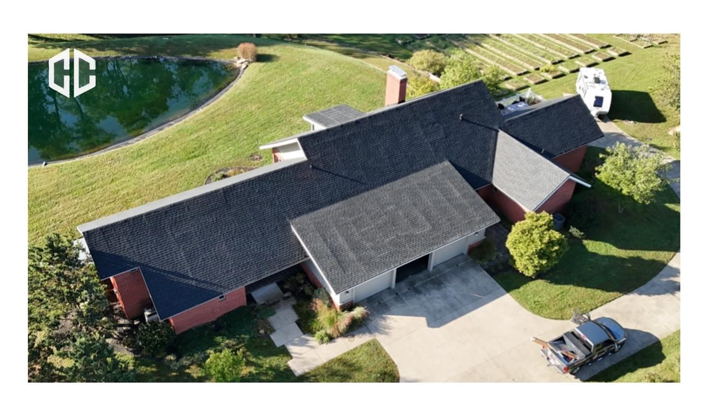 Aerial view of a house with a dark asphalt shingle roof, a pond, and a driveway with a truck and camper.