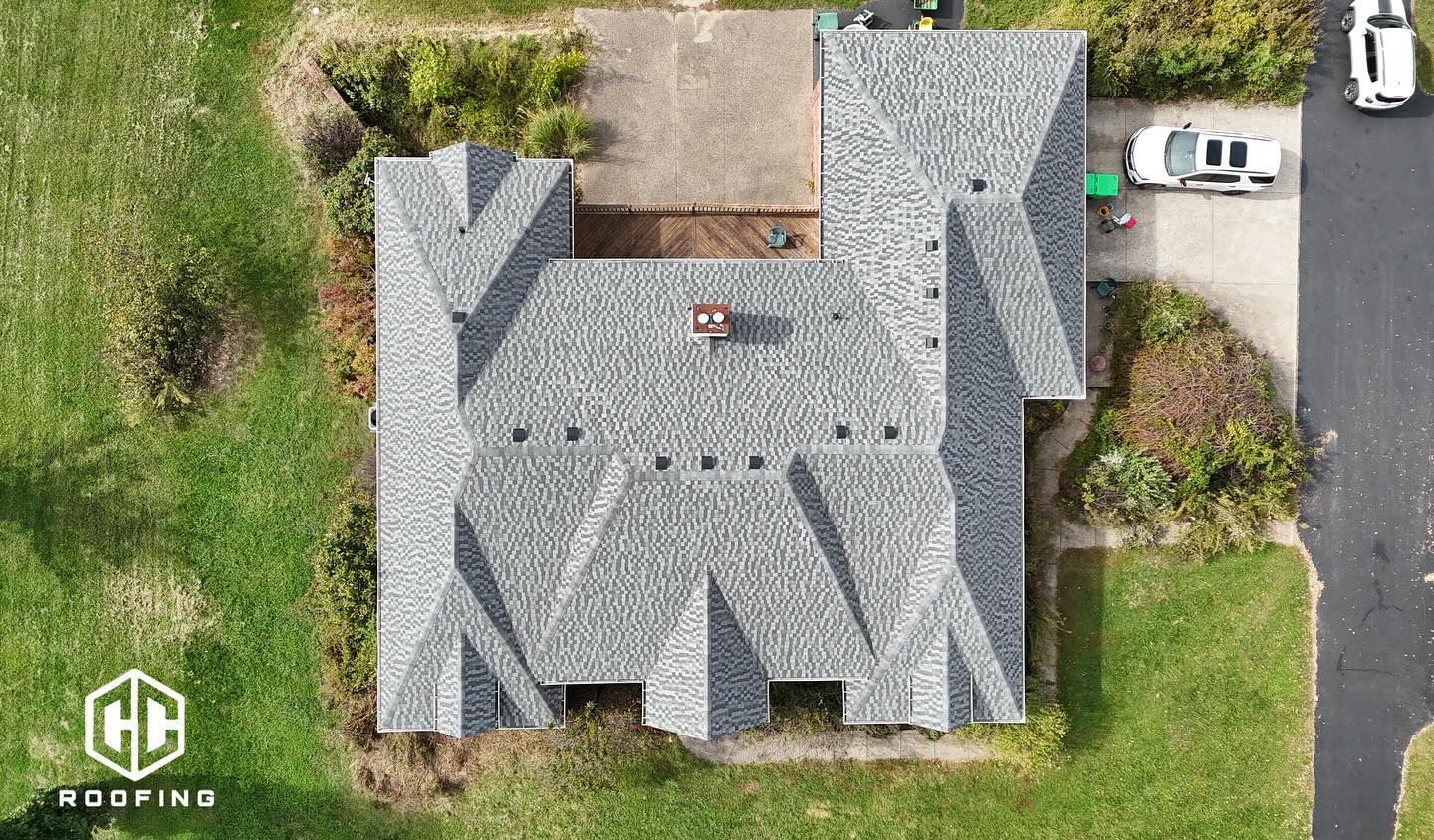 Aerial view of a residential house with a complex grey asphalt shingle roof, surrounded by lawn and a driveway.