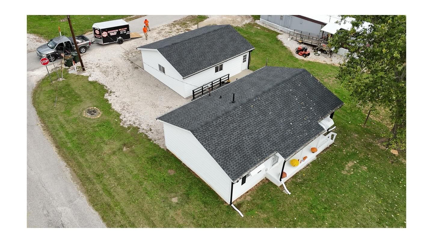 Aerial view of two white buildings with dark gray roofs, surrounded by gravel, green grass, and trees.