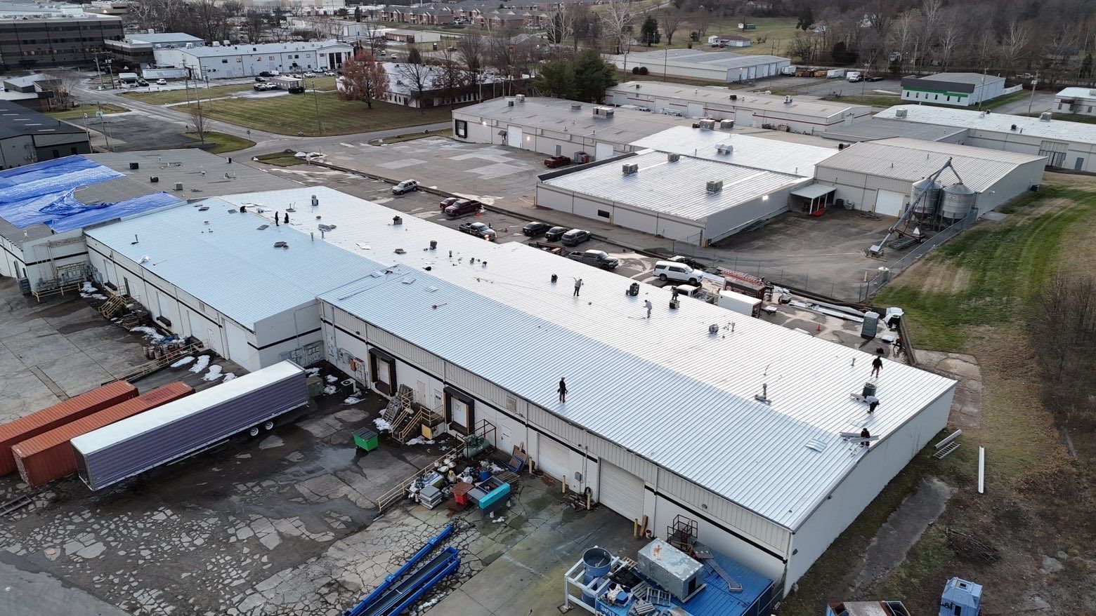 Aerial view of a long white commercial building with loading docks, parked semi-trailers, and surrounding industrial area.