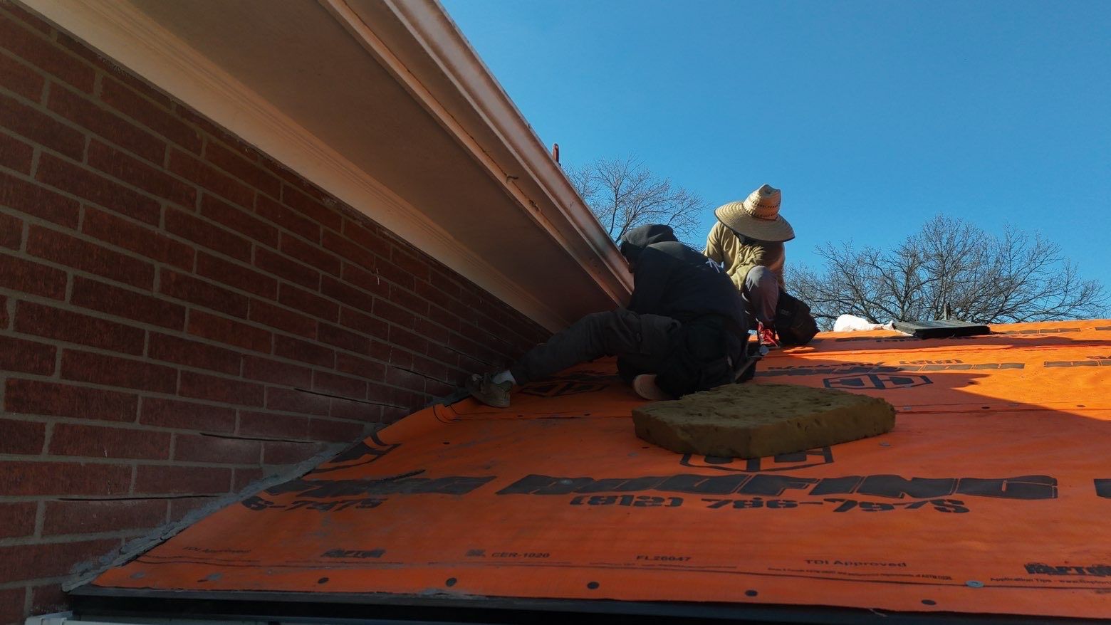 Two roofers wearing protective gear work on a residential roof covered in orange underlayment near a brick wall.