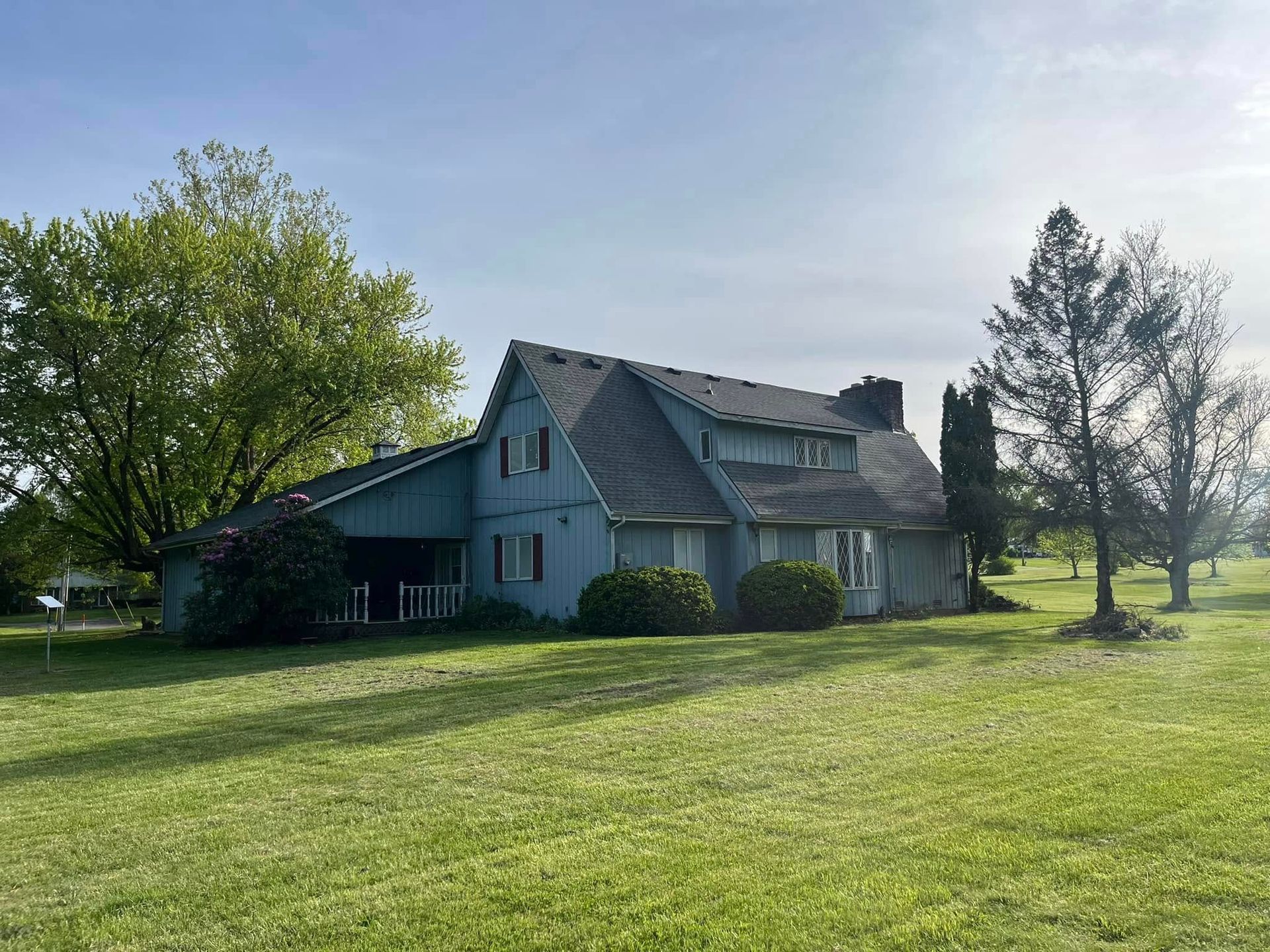 A two-story blue house with a dark gabled roof sits on a grassy field under a bright, clear sky.