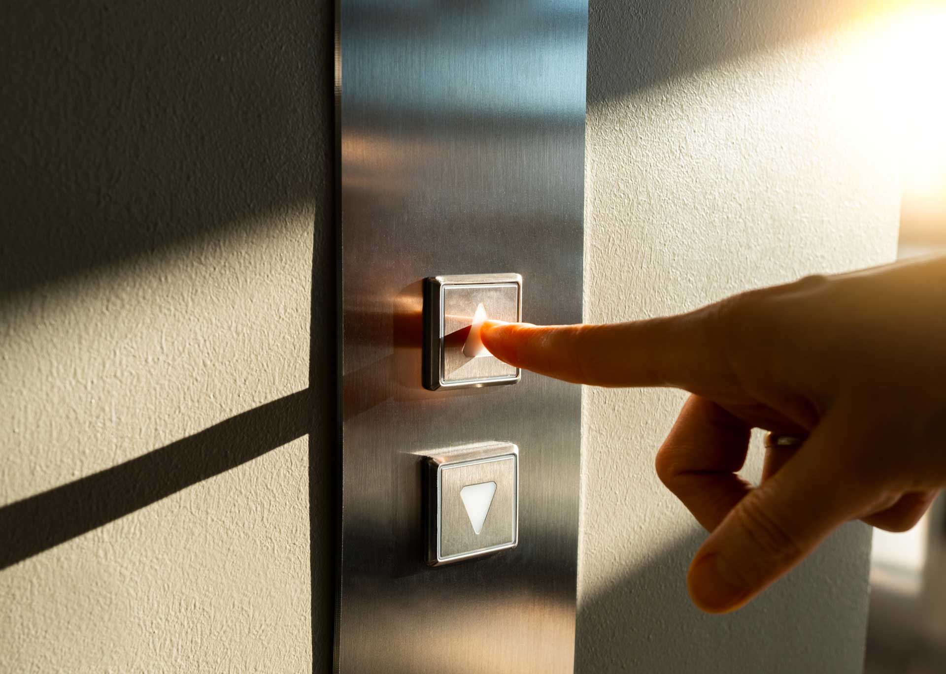 Hand pressing an illuminated elevator button on a brushed metal panel, in sunlight.