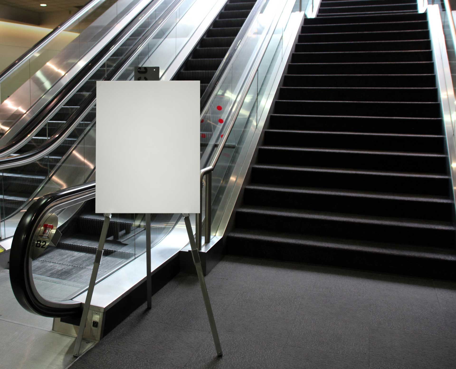 White blank sign on a metal easel in front of escalators and stairs in a building.