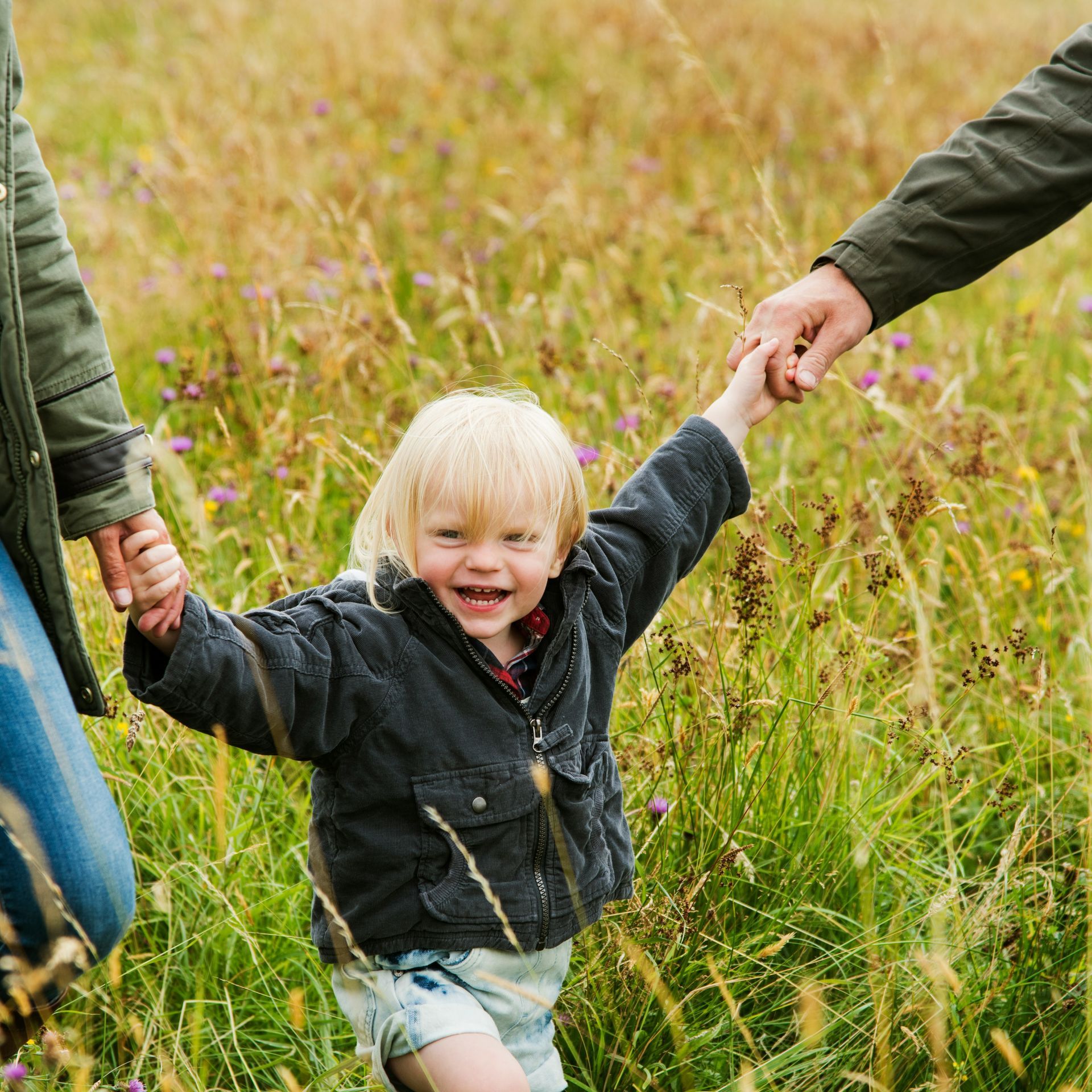 A little boy is holding the hands of his parents in a field.