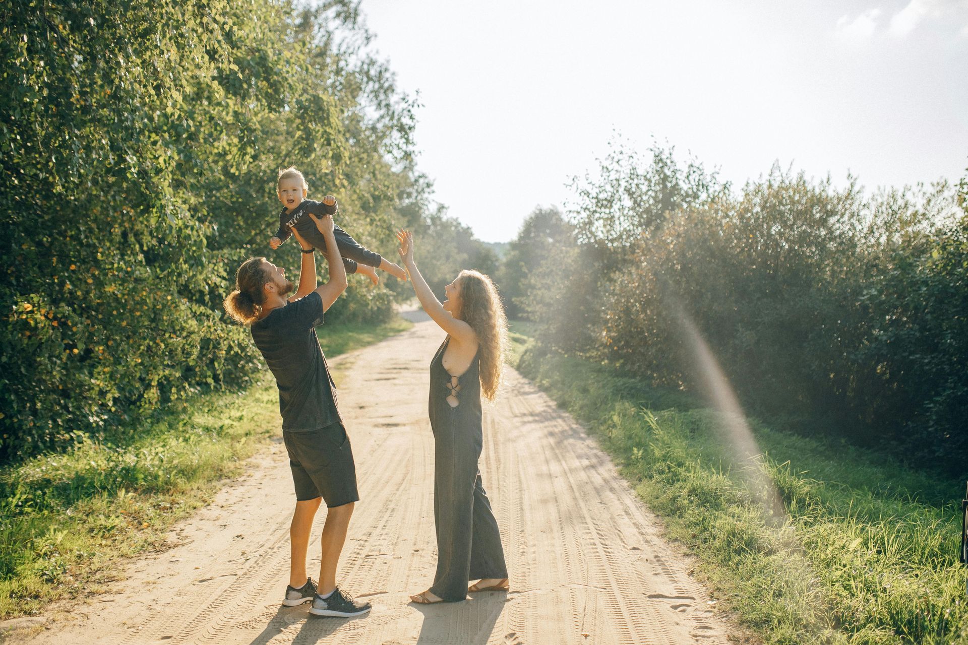 A man and woman are holding a baby in the air on a dirt road.