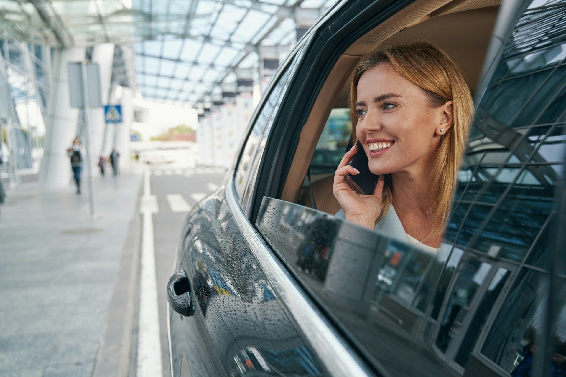Woman in car talking on phone near airport, smiling.
