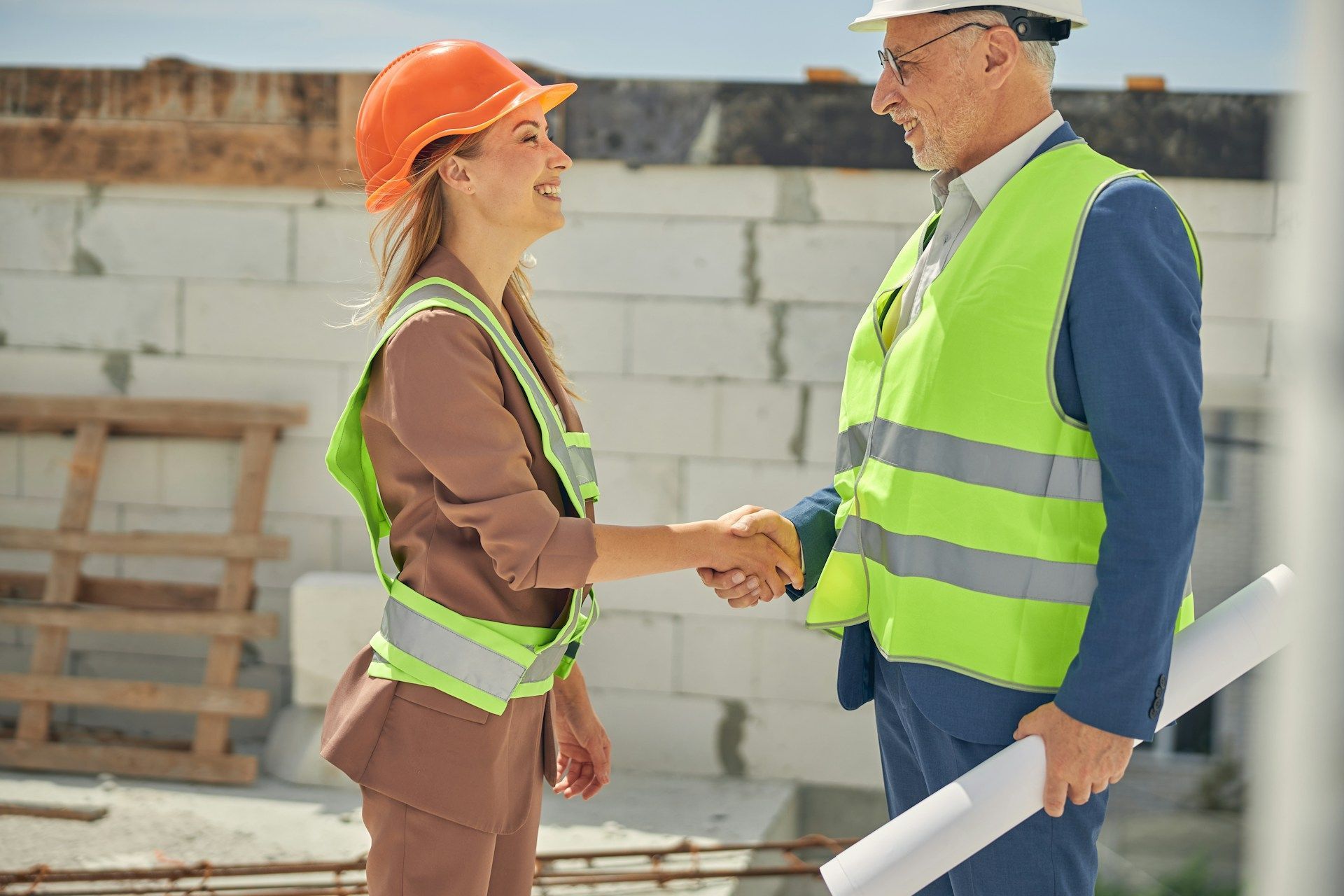 Woman in orange hard hat shakes hands with man in white hard hat on construction site. Both wear safety vests.