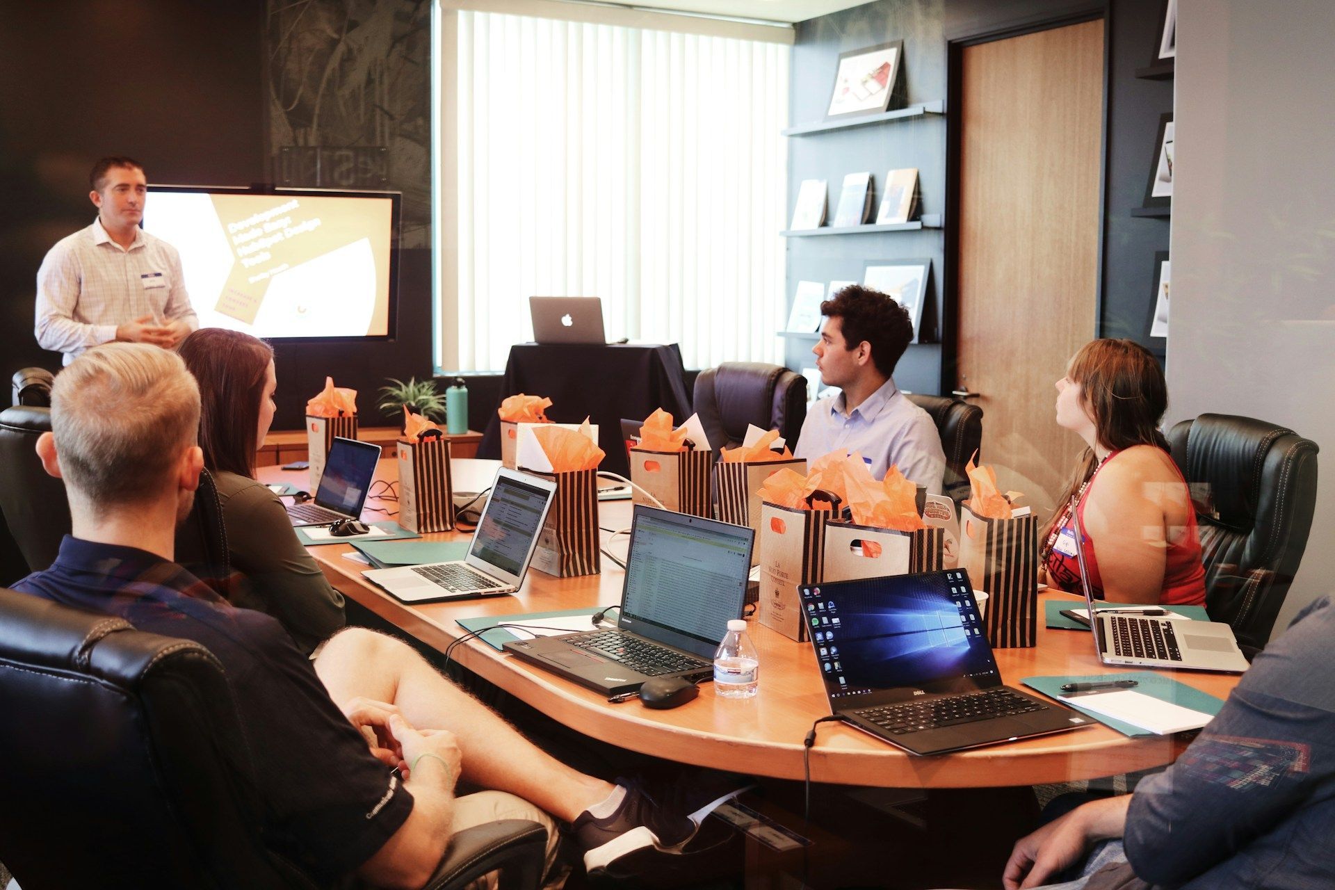 People in a meeting room, a man presenting to the group. Laptops and gifts on the table.