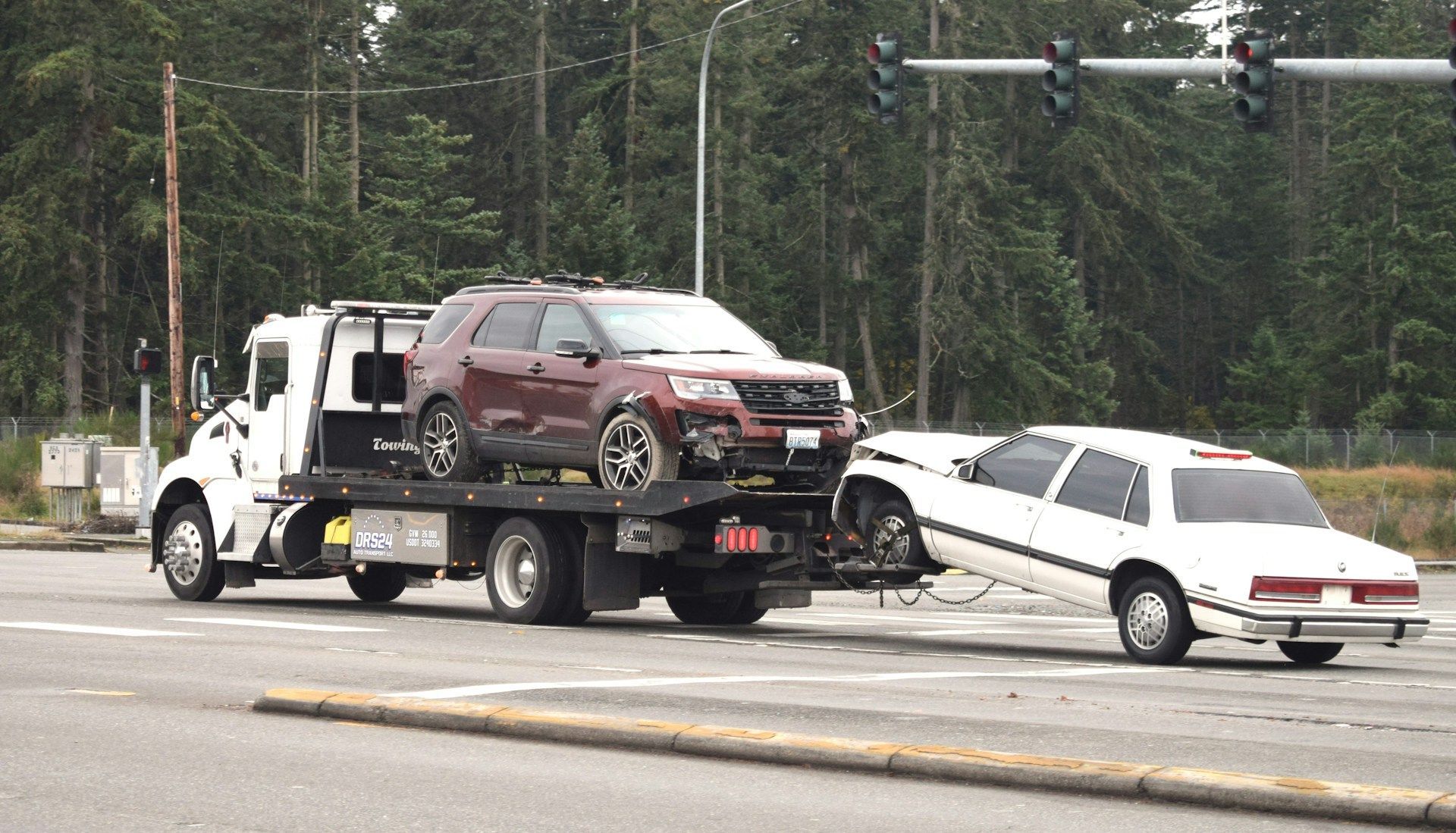 Tow truck with two damaged vehicles, a red SUV and a white sedan, at a traffic intersection.