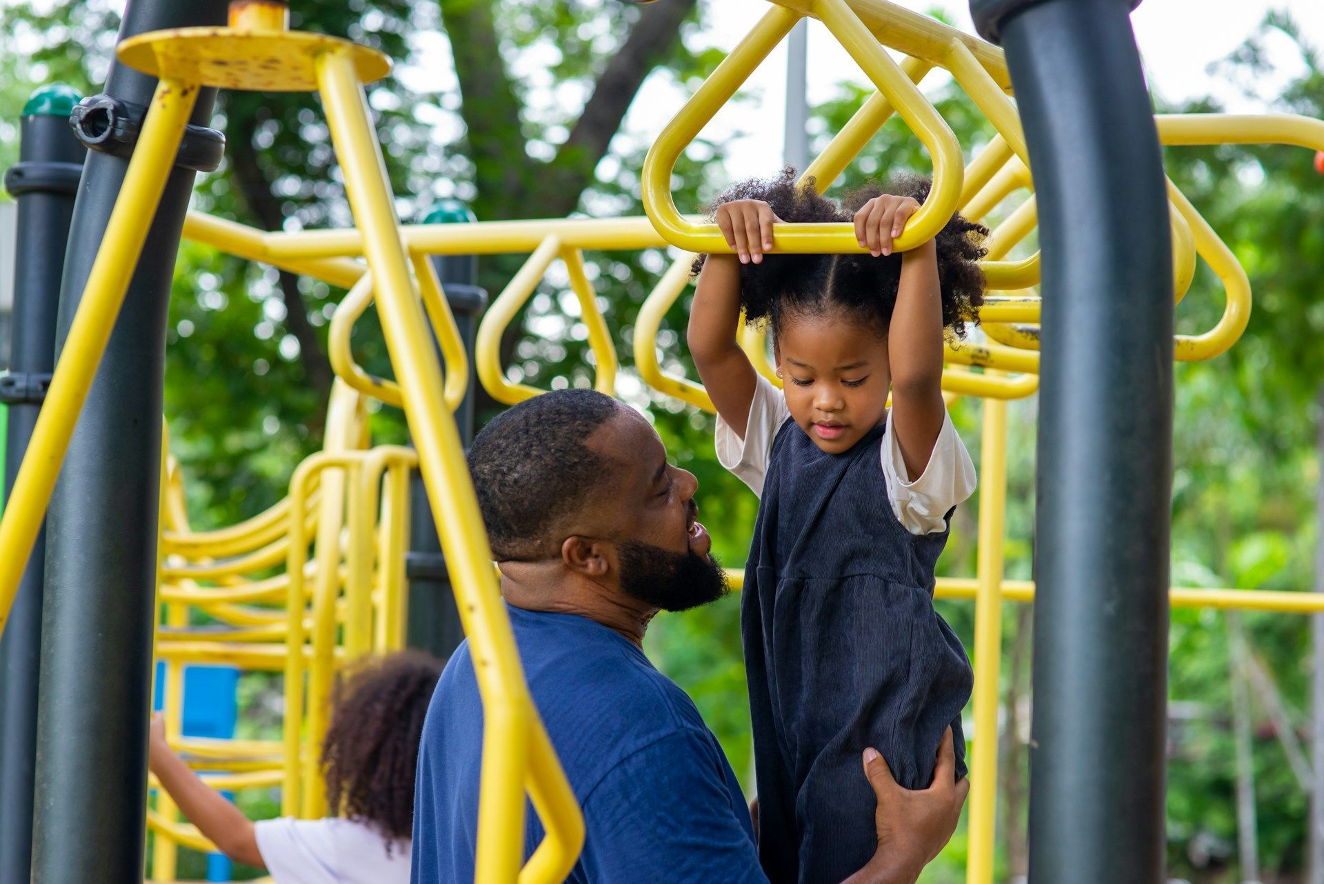 Father helps child on yellow monkey bars at a playground.