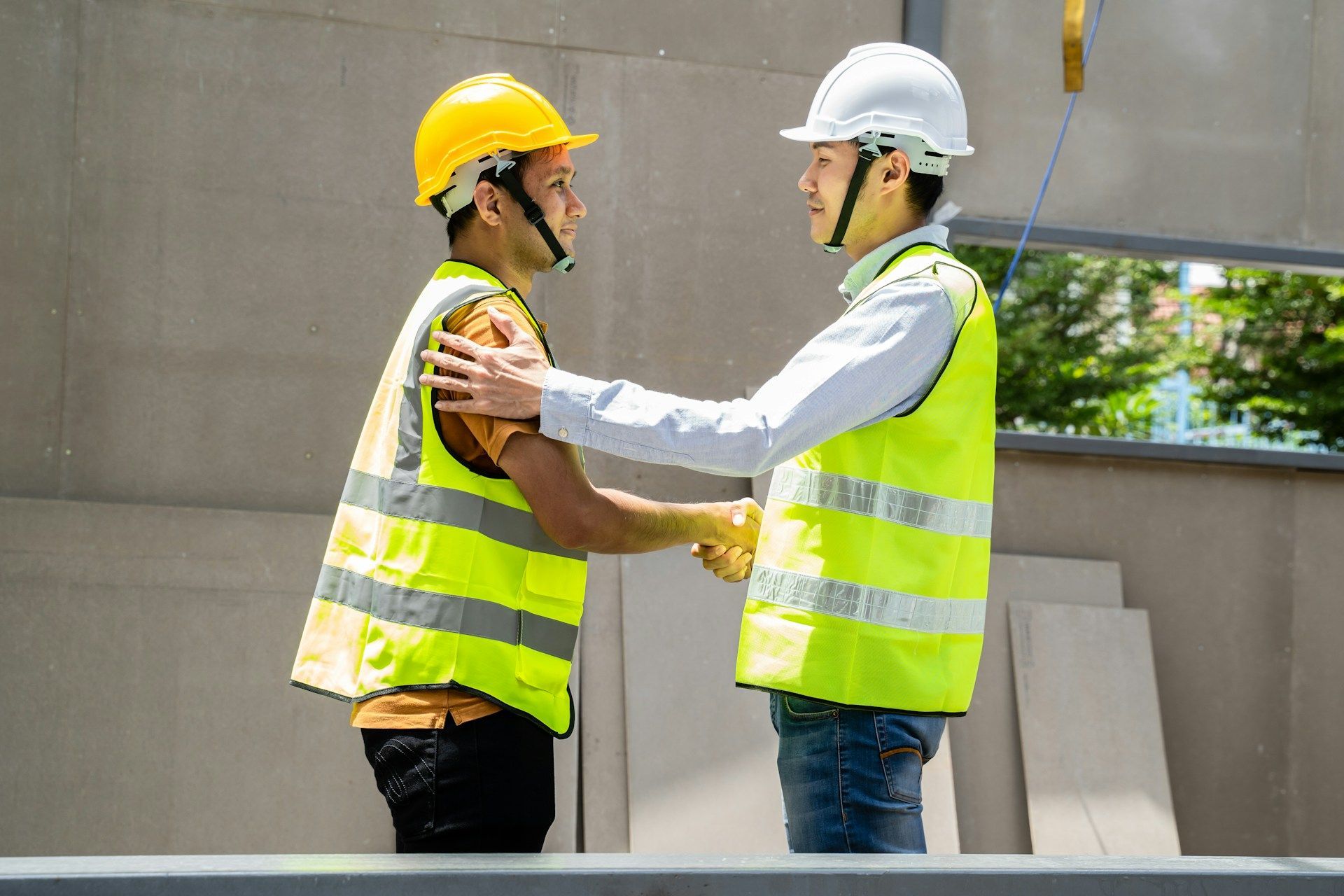 Two construction workers in safety vests and hard hats shake hands at a building site.
