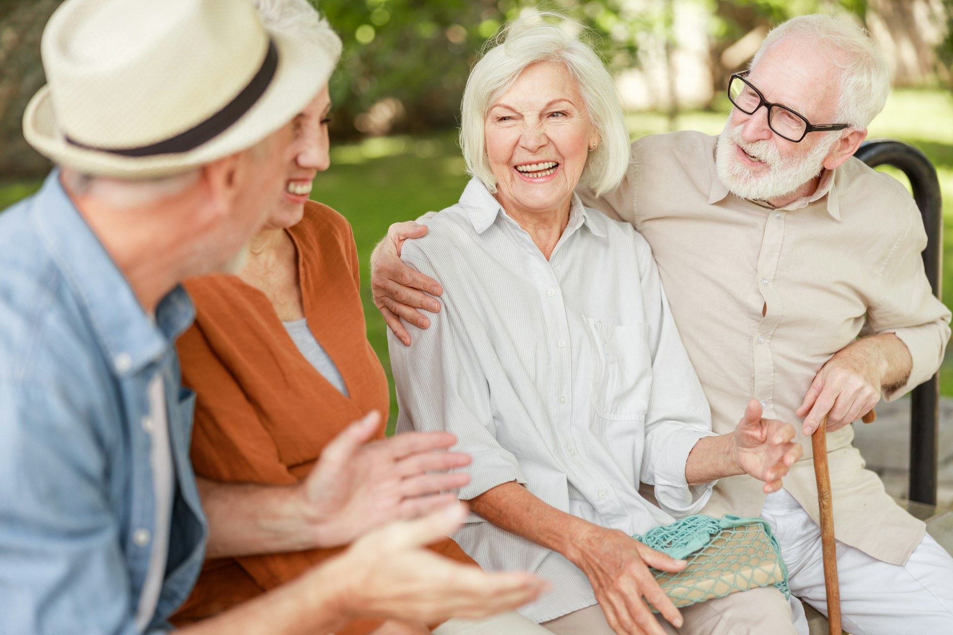 Four older adults laugh together on a park bench. Man with cane has arm around woman.