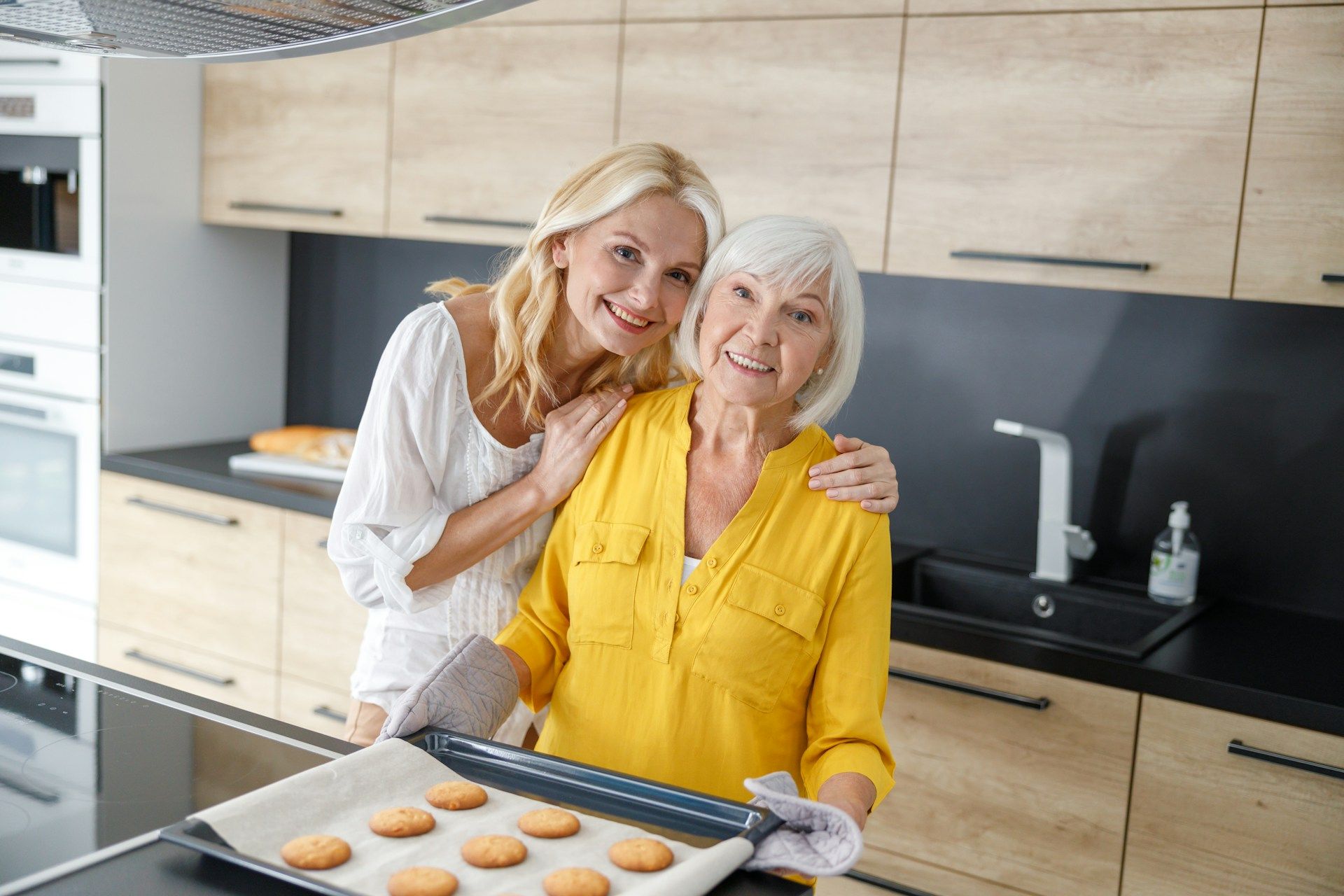 Woman and older person with cookies in kitchen. Woman embraces older person holding baking sheet.