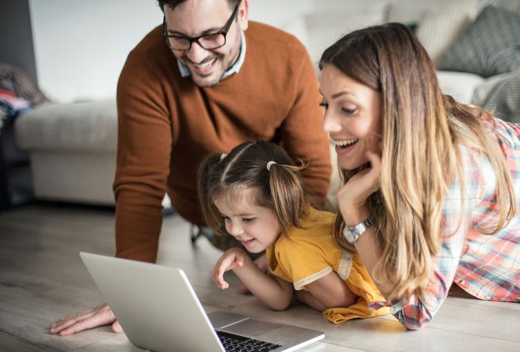 Family of three looking at a laptop on the floor; smiling faces and neutral indoor setting.