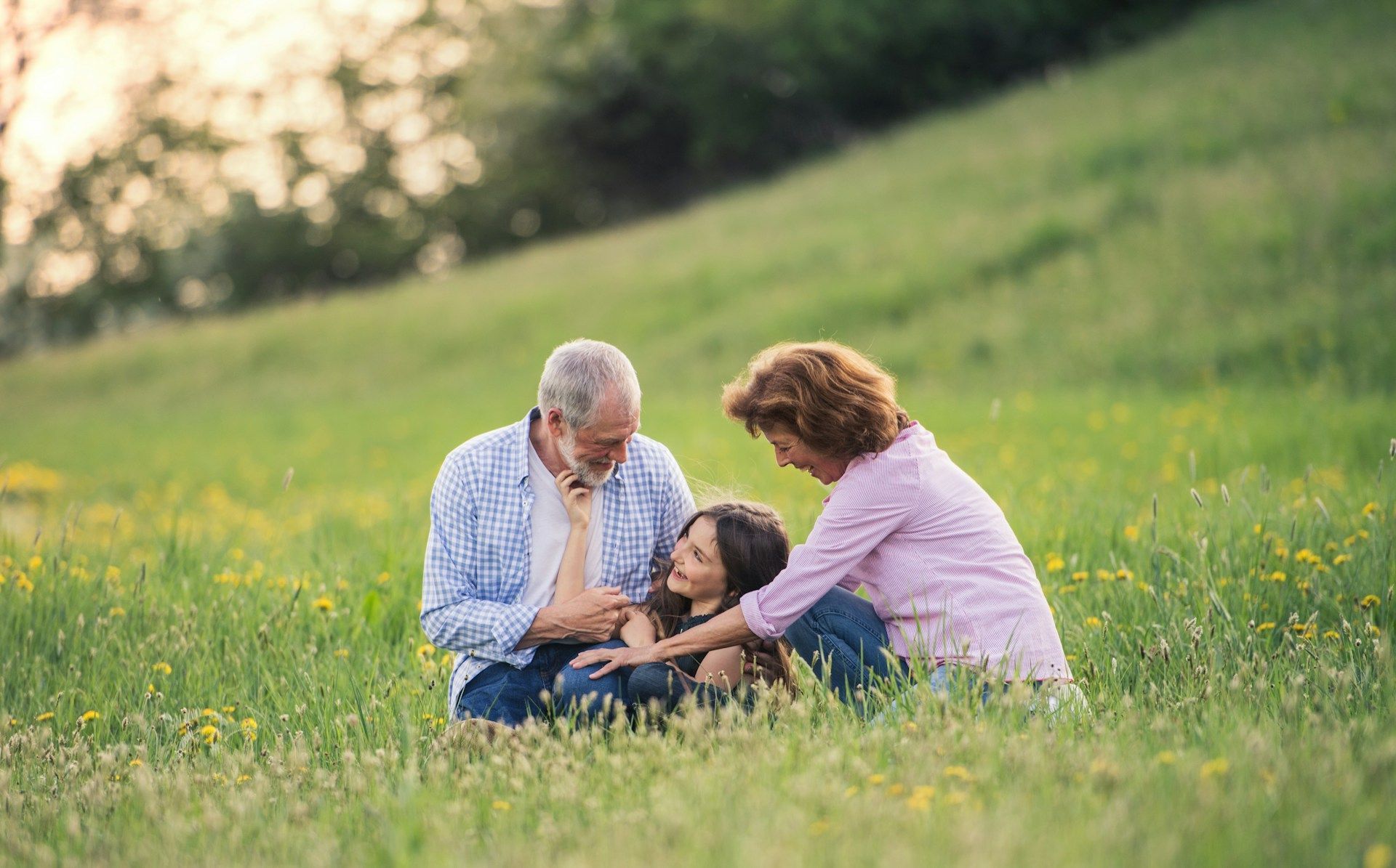 Grandparents and grandchild kneeling in a grassy field, looking at something together.
