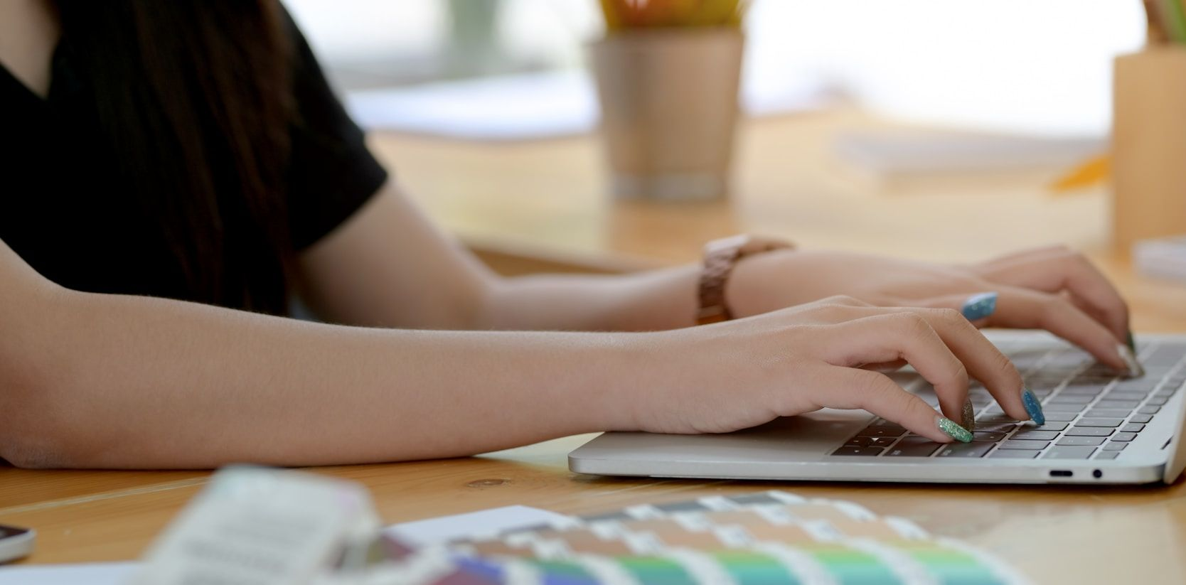 Person typing on a laptop at a wooden desk with color swatches and office supplies.