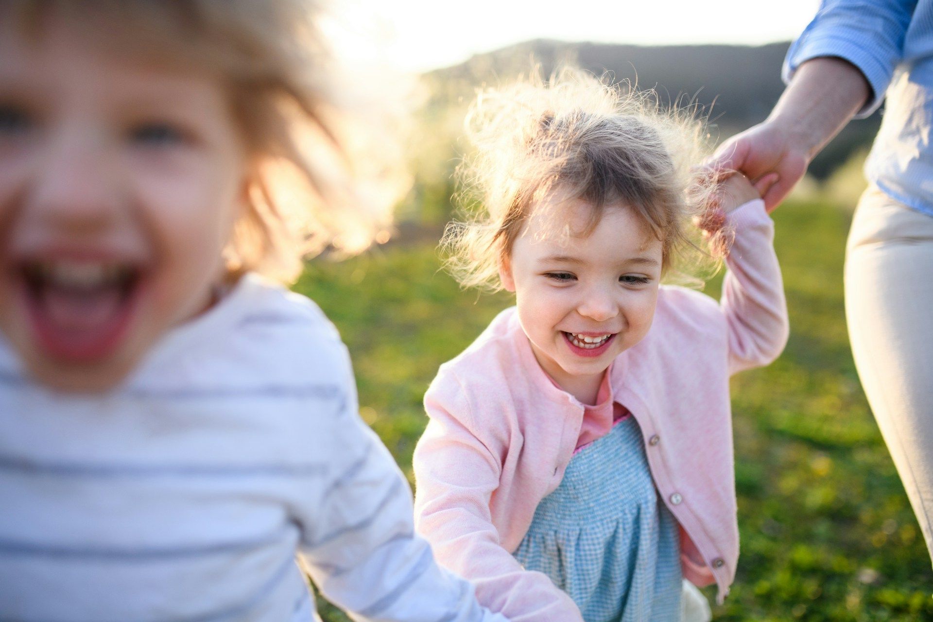 Two smiling children run in a grassy field, one held by an adult's hand. Warm sunlight.