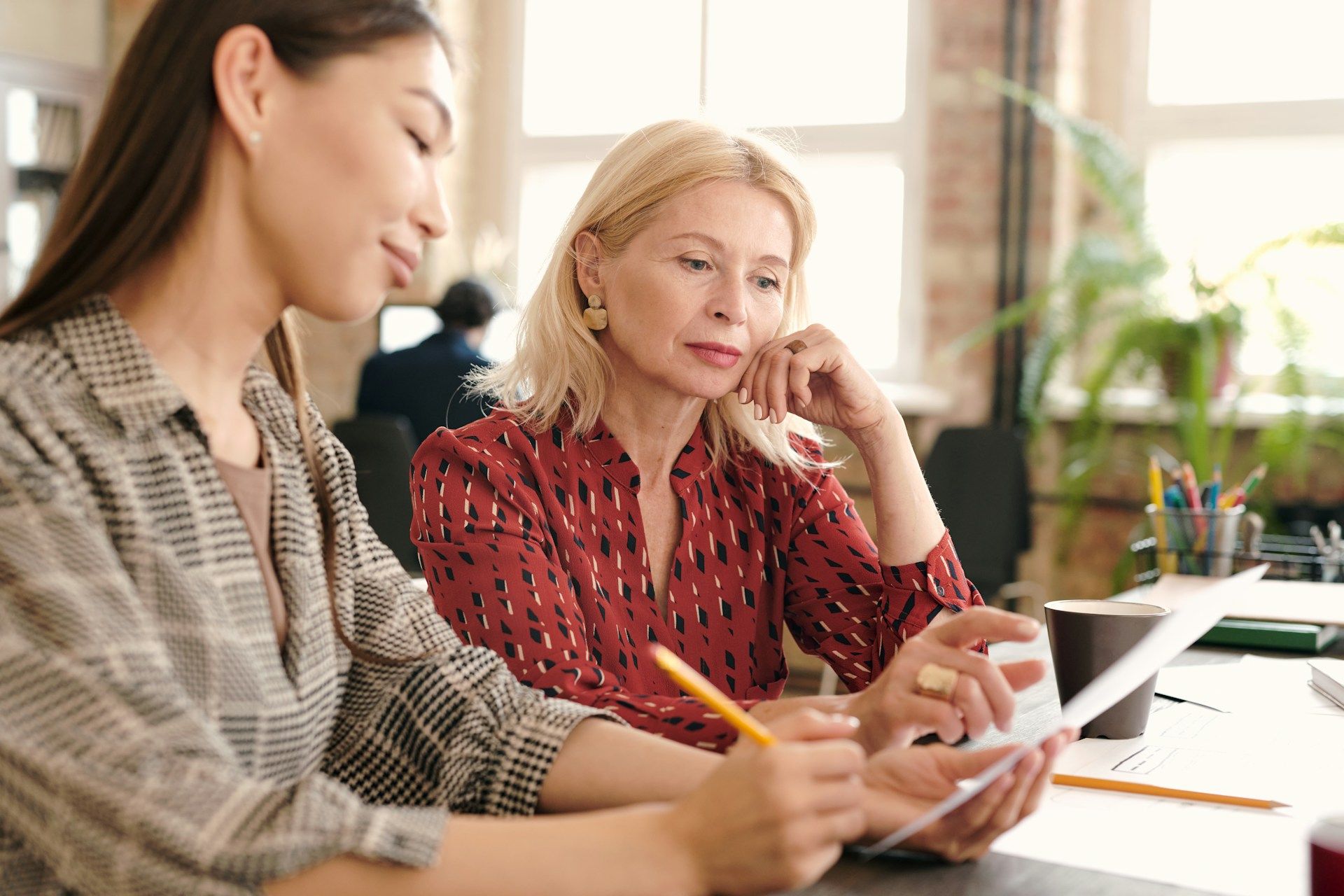 Two women reviewing documents at a desk in an office setting. One points with a pen, the other looks on.