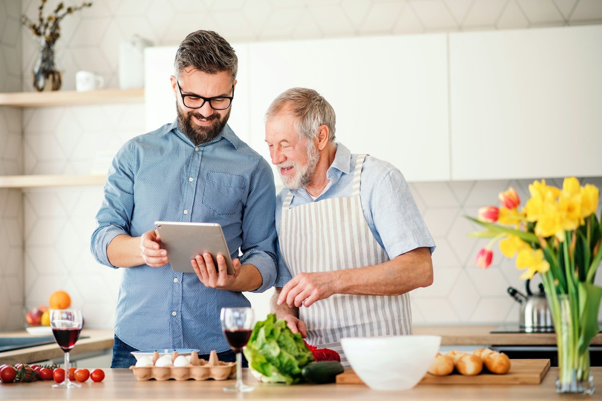 Man shows older man tablet while cooking in kitchen.