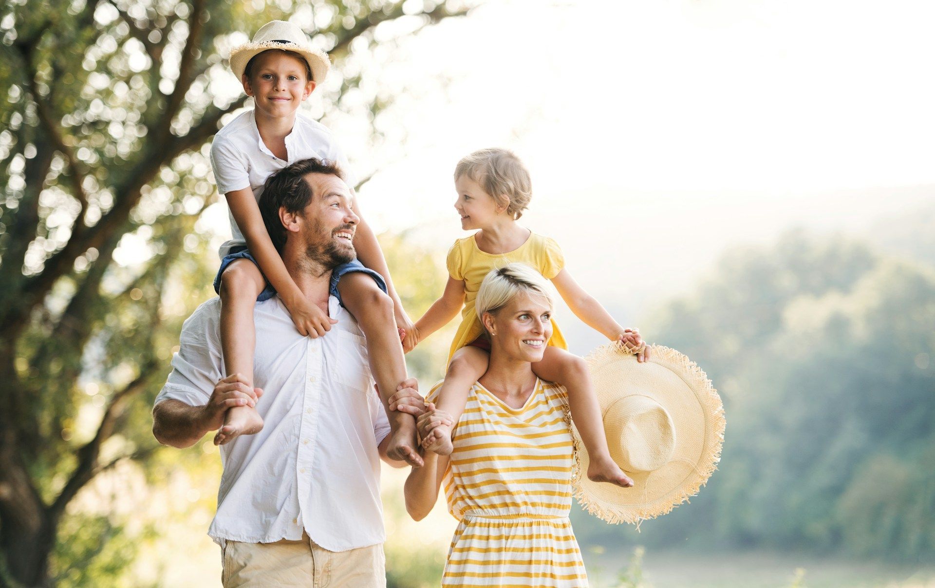 Family with two children on their shoulders, smiling outdoors, sunny setting.