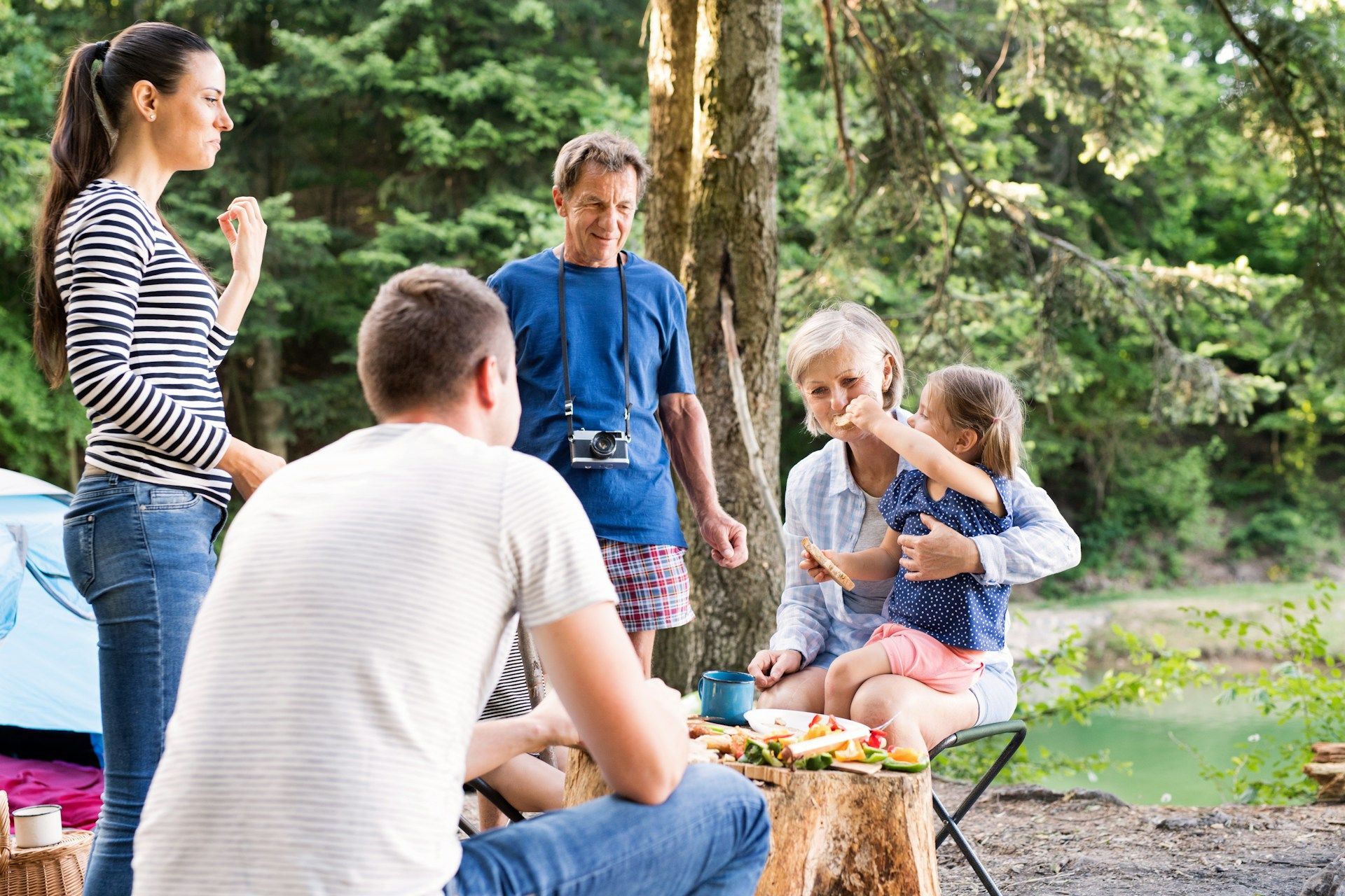 Group of people outdoors, gathered near a tree. One person holds a child. Camping, casual setting.