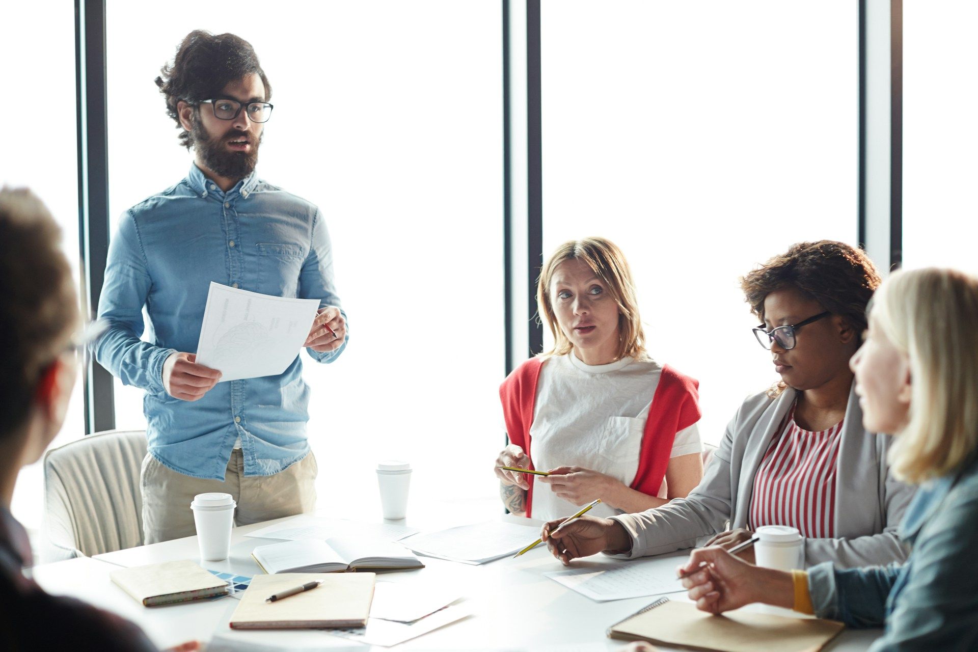 Man presenting to a group in a brightly lit meeting room. People seated around a table, engaged.